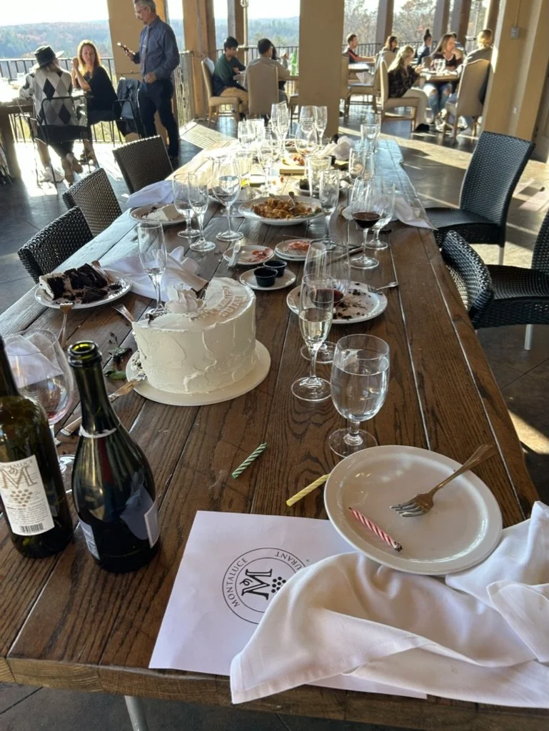 A wooden table set for a celebration with empty wine glasses, a birthday cake, leftover cake slices, bottles of wine or champagne, and some plates with crumbs and a birthday candle. In the background, people are dining indoors and outdoors, enjoying 