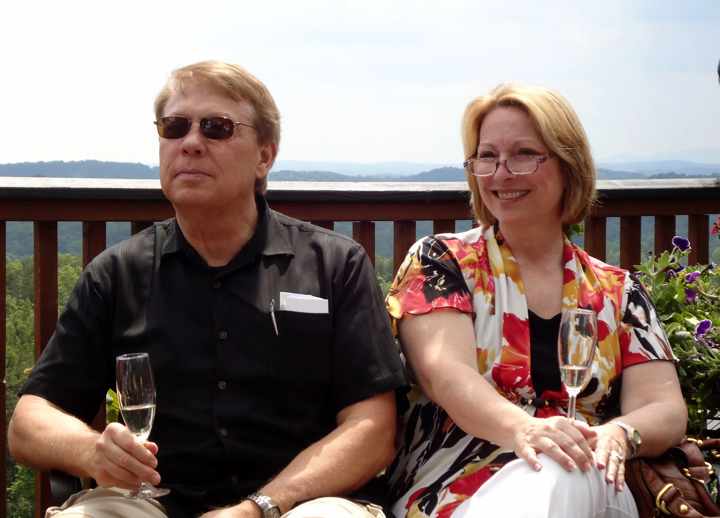 A man and woman sitting outdoors on a deck with a scenic view of mountains and trees in the background, holding glasses of champagne, smiling and enjoying a sunny day.
