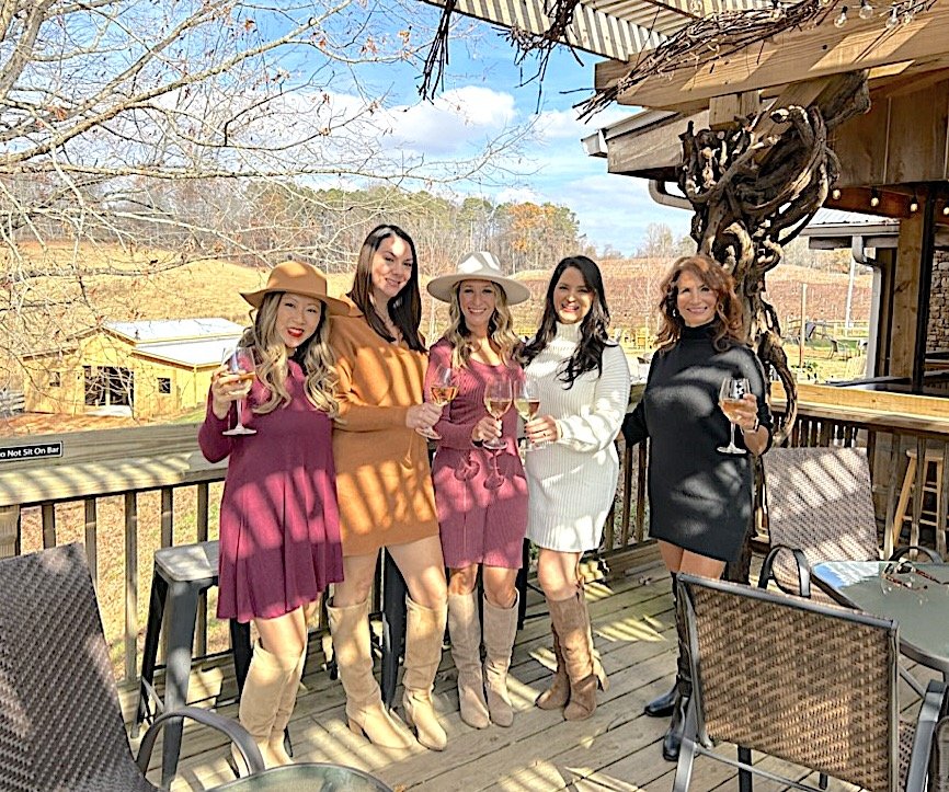 Five women standing on a wooden deck outdoors, holding glasses of wine, smiling and dressed in stylish fall outfits with hats and boots, with a scenic rural landscape in the background.