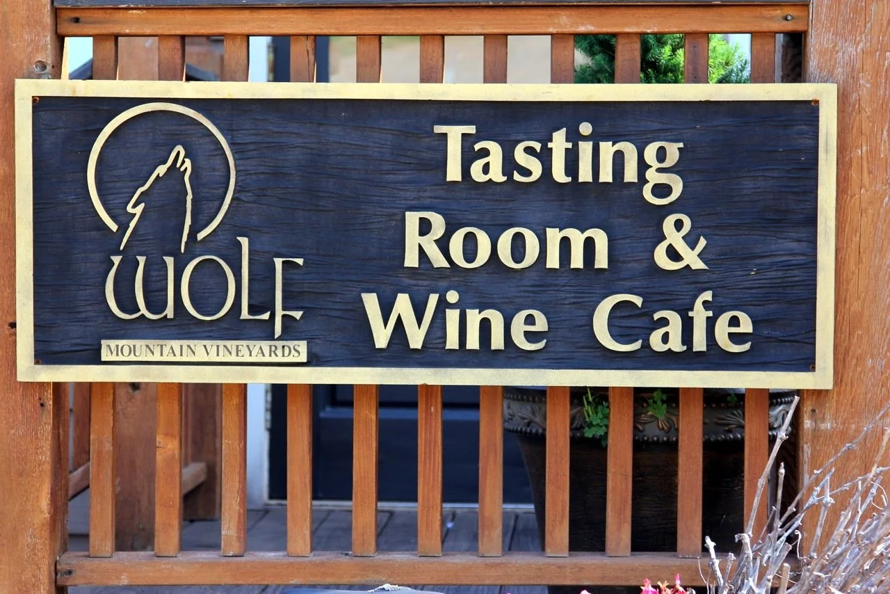 A wooden sign for Wolf Mountain Vineyards indicating a tasting room and wine cafe. The sign features a stylized wolf howling inside a circle and the words "Tasting Room & Wine Cafe" in large, light-colored text on a dark background, mounted on a wood