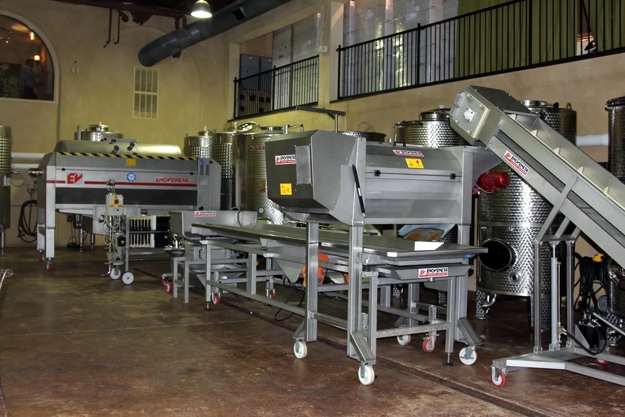 Industrial wine production equipment inside a winery, featuring stainless steel tanks, conveyors, and machinery used for processing grapes and making wine.