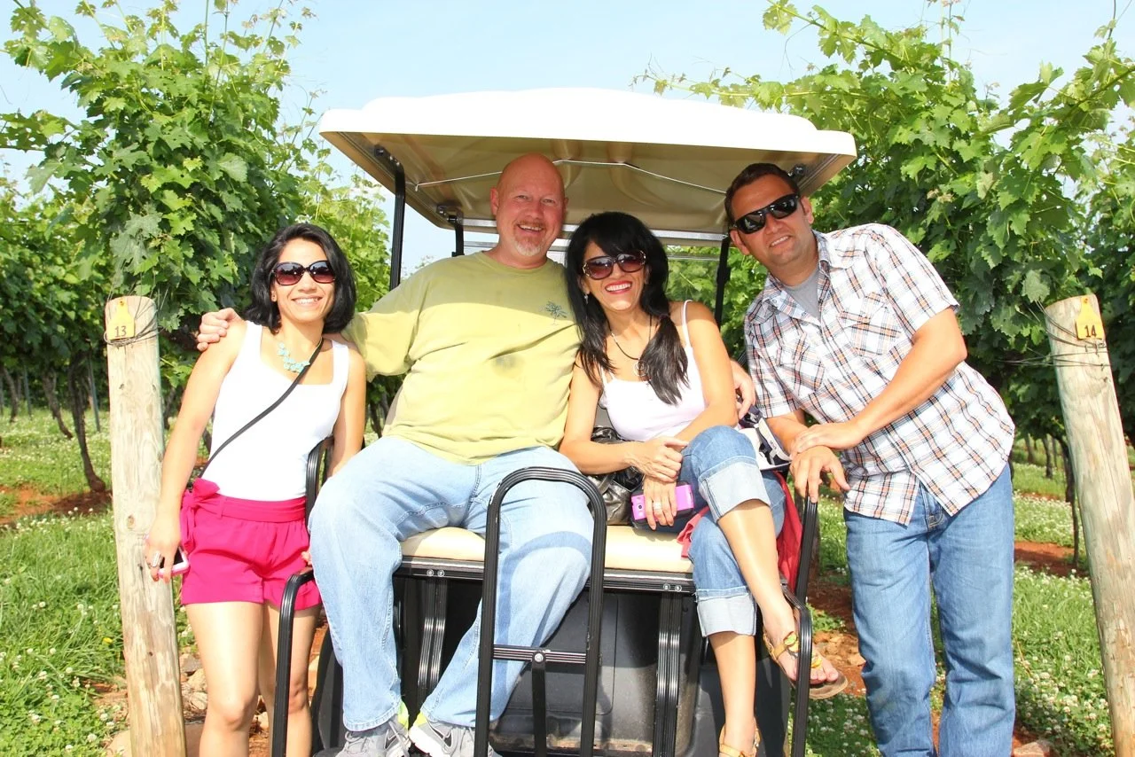 Four people smiling and sitting on a golf cart in a vineyard, enjoying a sunny day.