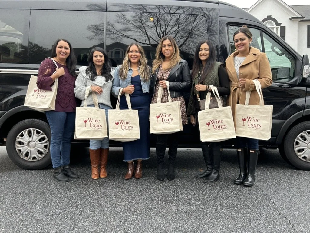 Six women standing in front of a black van, each holding a beige tote bag with the words 'Wine Tours' and a wine glass logo printed on it. They are smiling and dressed in casual to semi-casual clothing.