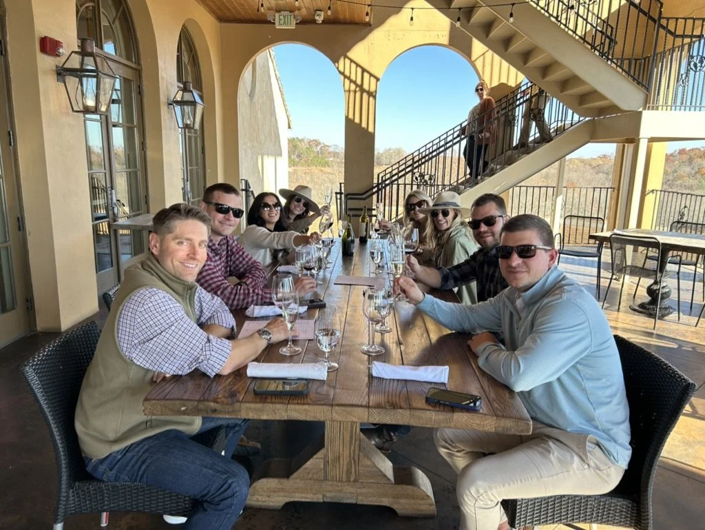 People having a meal at a long outdoor dining table with wine glasses, with a woman standing on a staircase in the background.