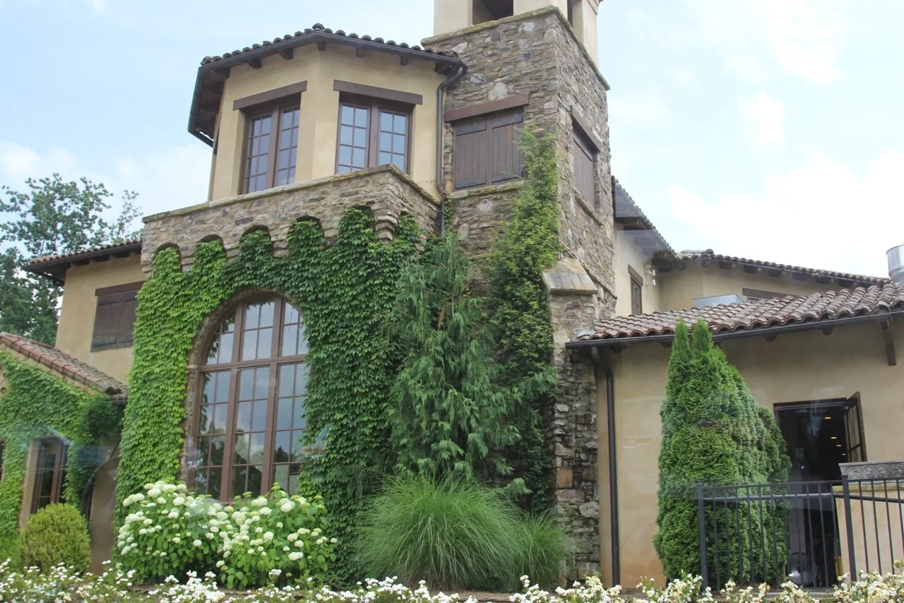 A large house with stone and stucco exterior, multiple windows, and a red tile roof surrounded by greenery and flowering plants.