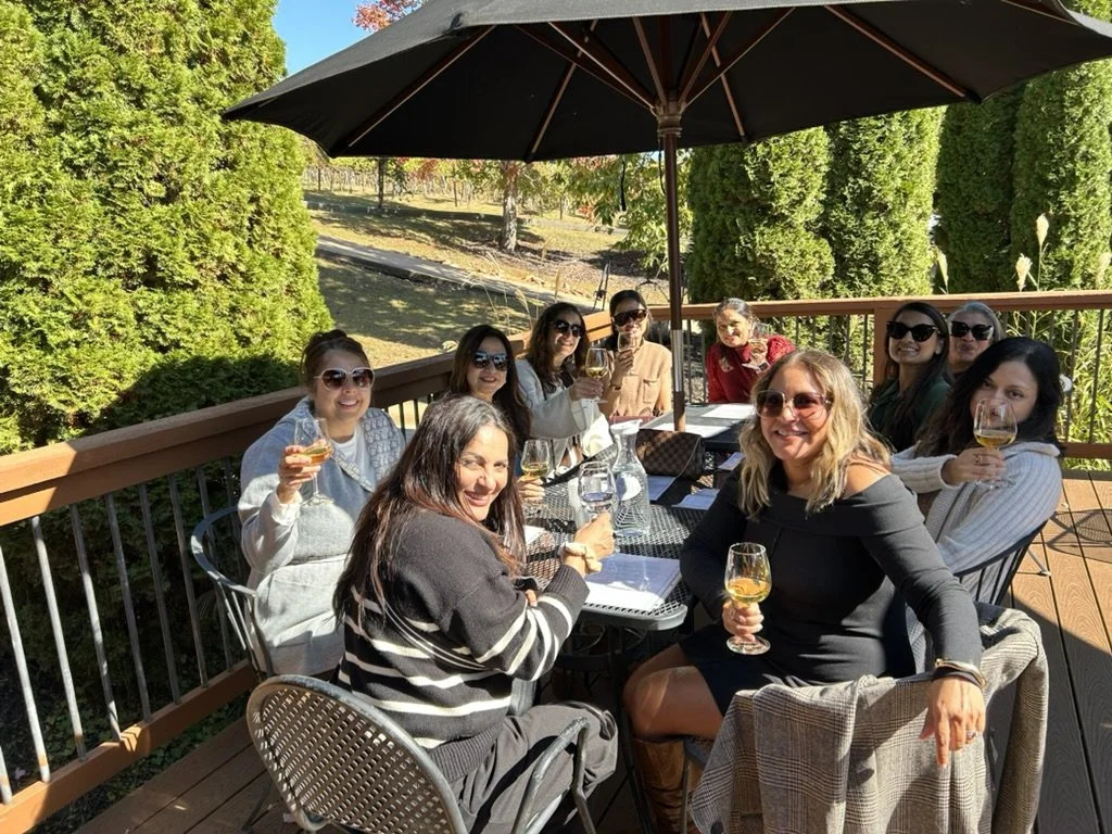 Group of women sitting on a balcony outdoors, enjoying drinks under a large umbrella, with greenery and trees in the background.