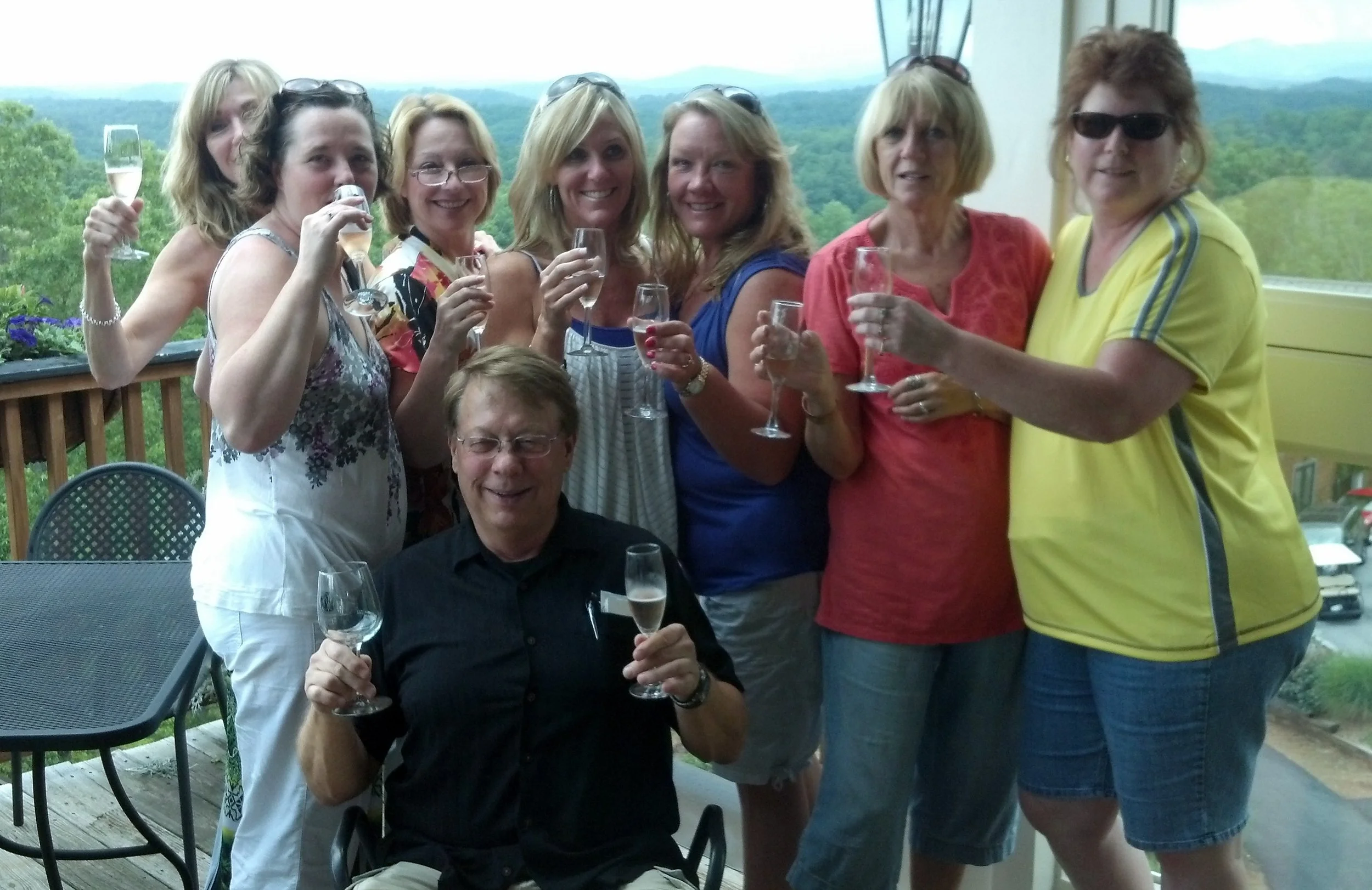 A group of nine people, mostly women and one man, celebrating with champagne glasses on a balcony overlooking a green landscape.