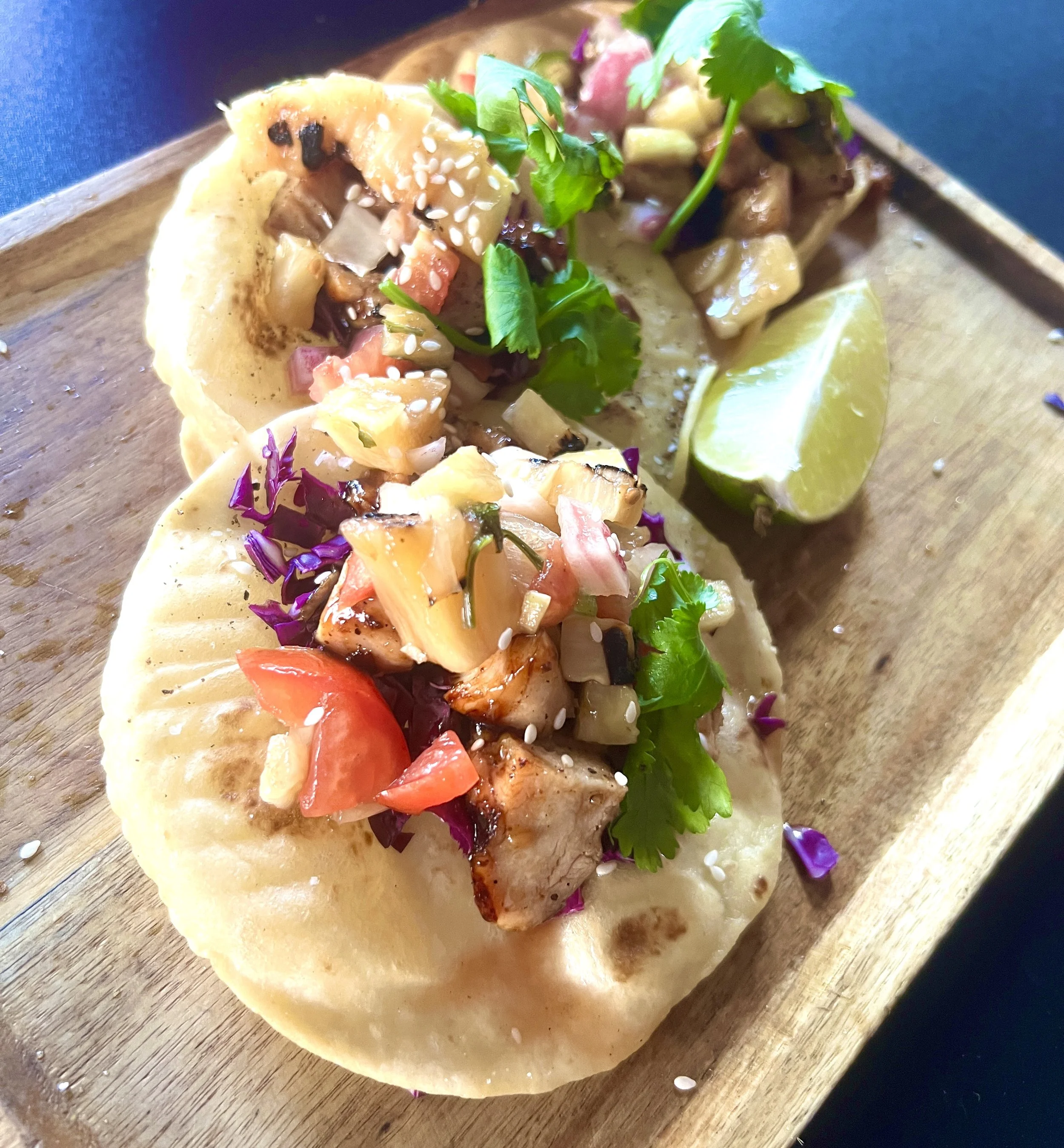 A taco with grilled chicken, diced tomatoes, chopped onions, cilantro, and sesame seeds in a soft corn tortilla on a wooden serving board, with a lime wedge.