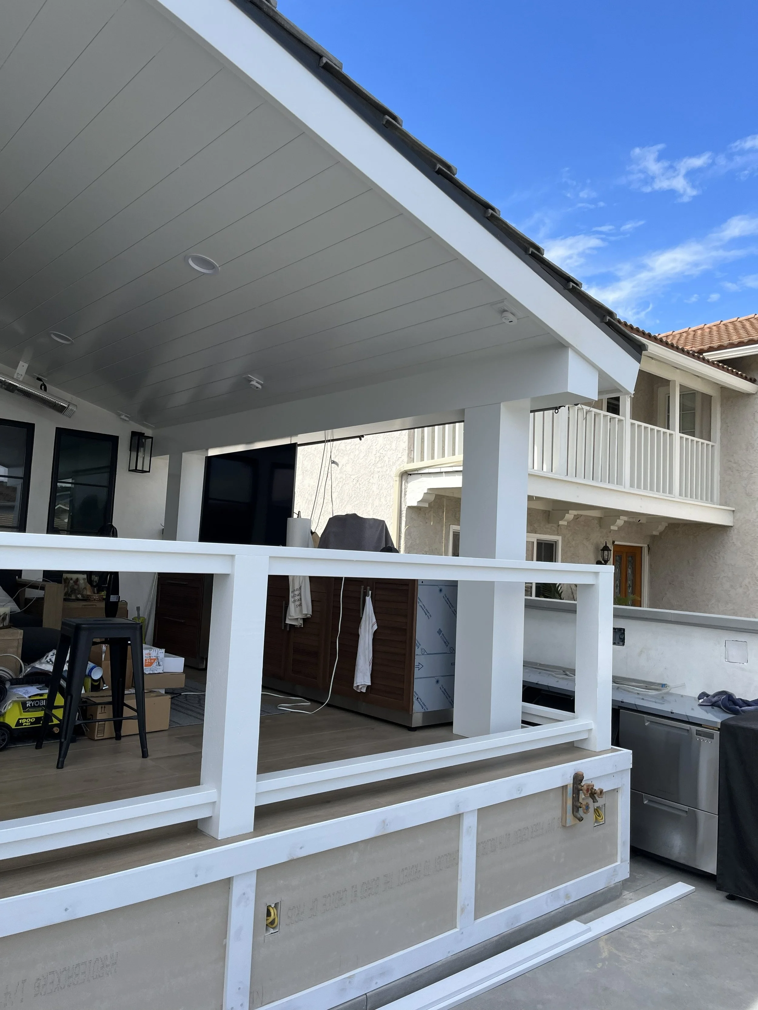 View of a partially constructed outdoor patio with white railing, ceiling, and support columns, under a blue sky.