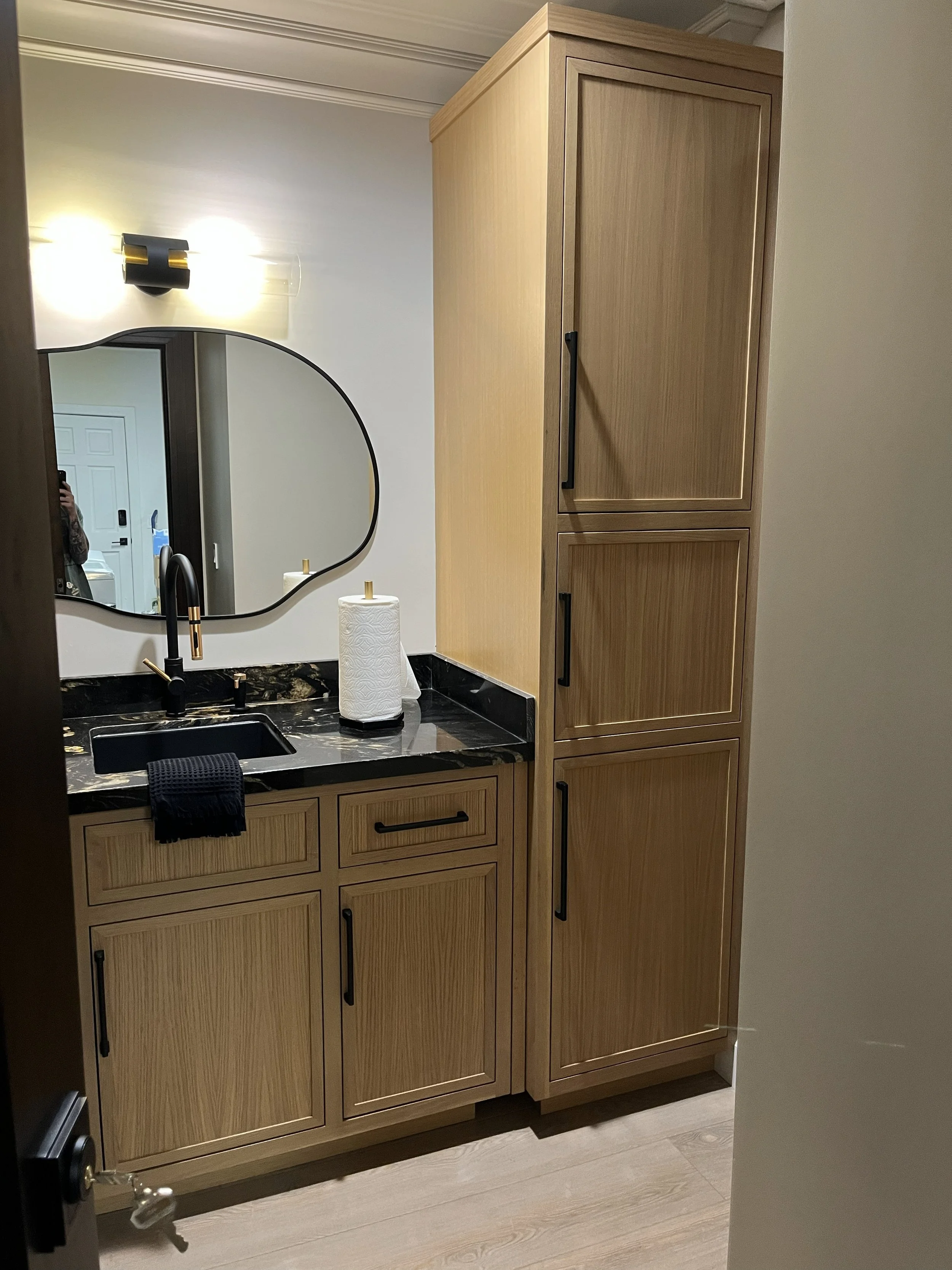 A bathroom vanity with a black marble countertop, a small black sink, a black faucet, a roll of paper towels, and a black towel hanging from the side. Above the vanity is an irregularly shaped mirror and a wall-mounted light fixture. To the right is a tall wooden cabinet and below the vanity are wooden cabinets with black handles.