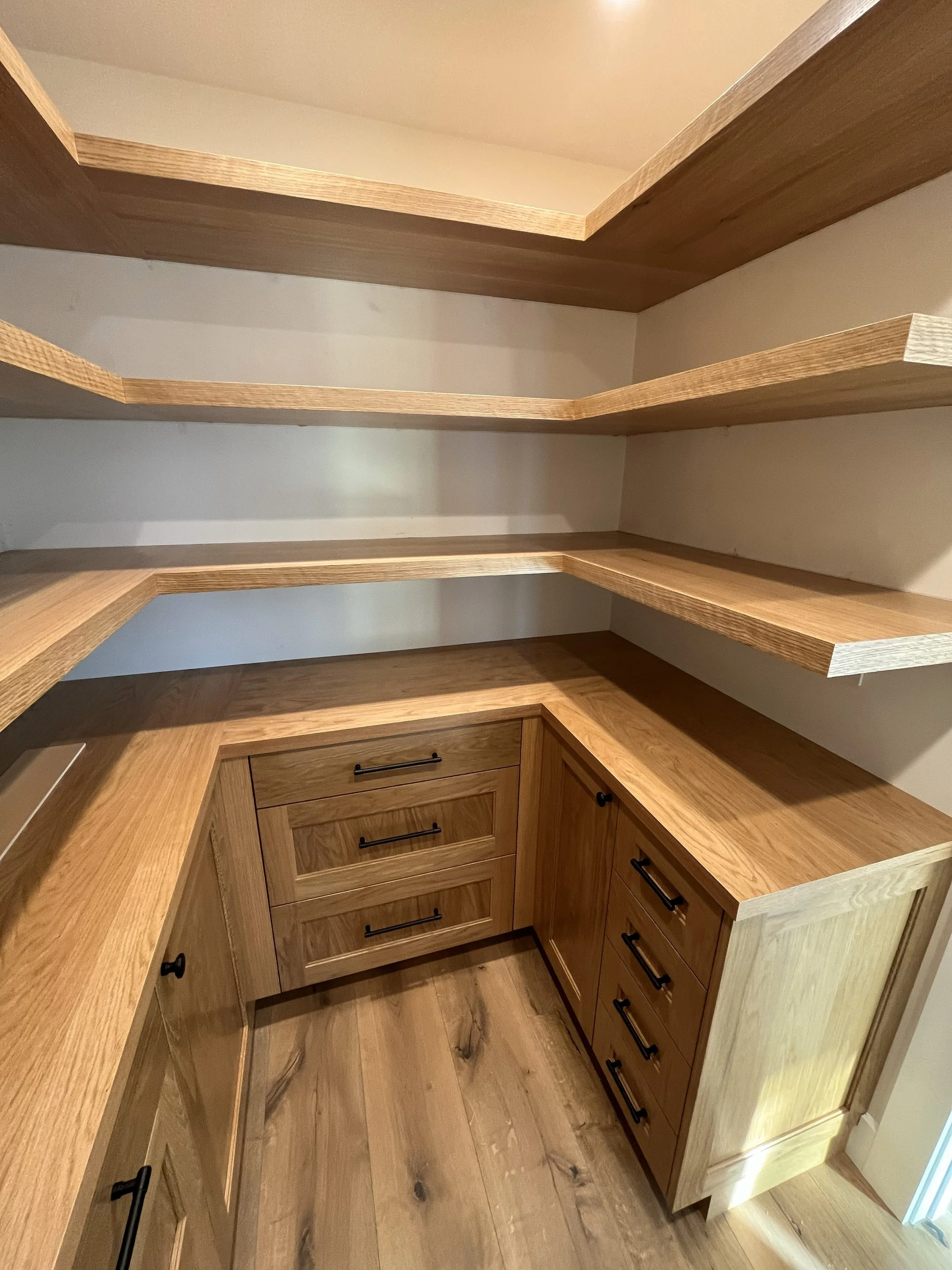 Empty corner of a custom wooden kitchen pantry with open shelves and lower cabinets with black handles.