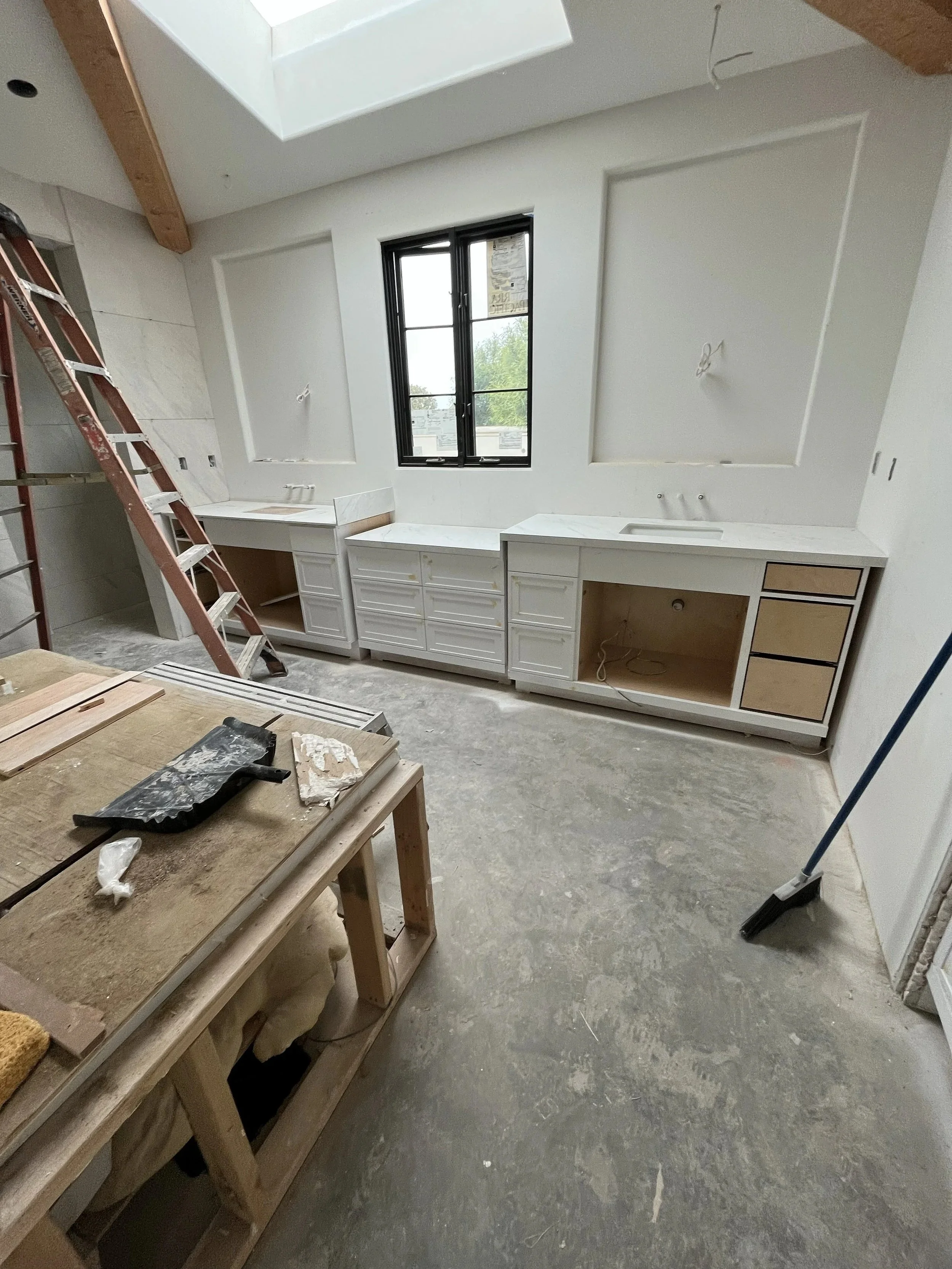 Kitchen under renovation with partially installed white cabinets, a large window, a ladder, construction tools on a wooden workbench, and unfinished flooring.