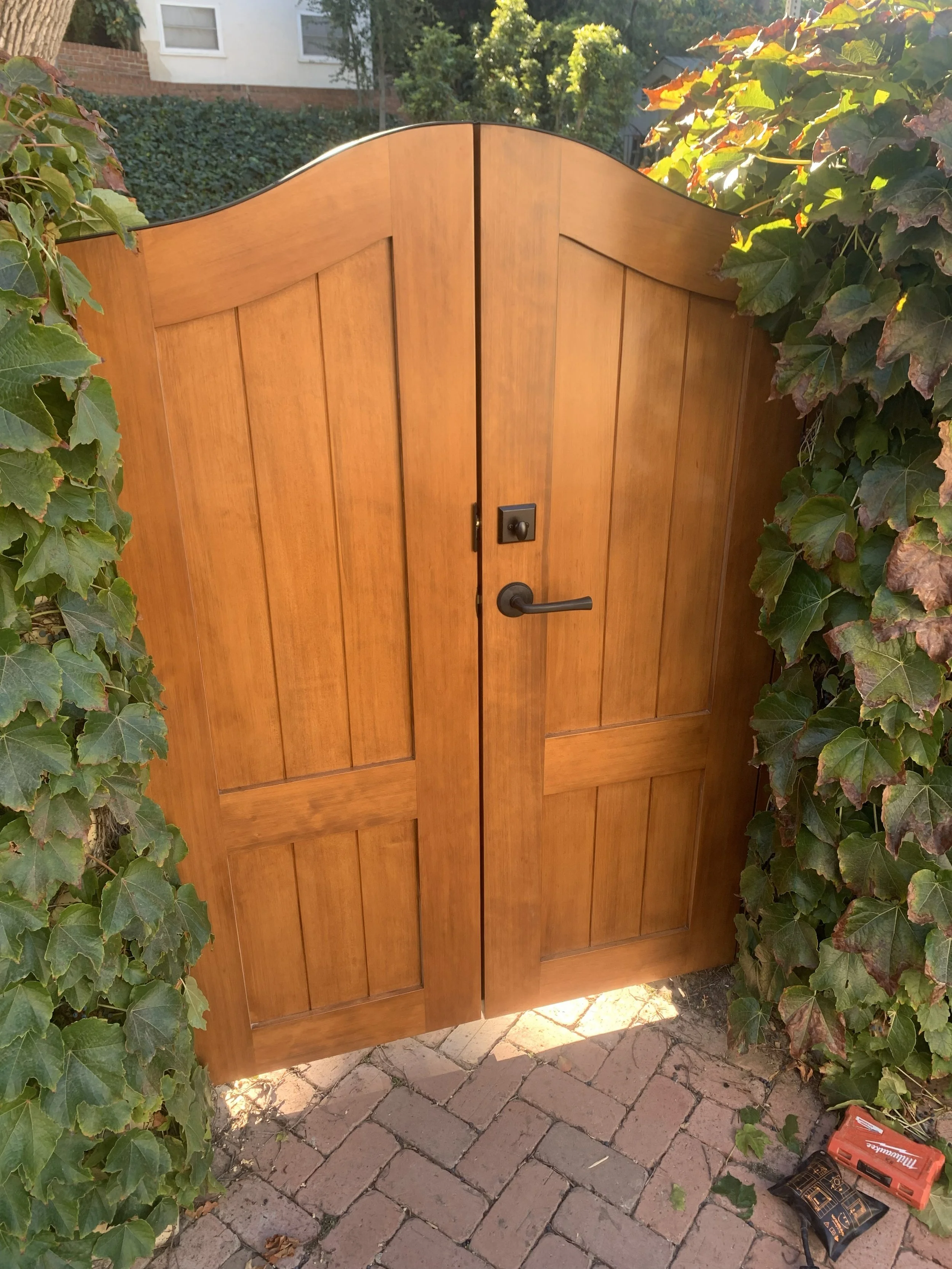 A wooden backyard gate with a black handle and lock, surrounded by green ivy and plants, with a brick pathway in front.