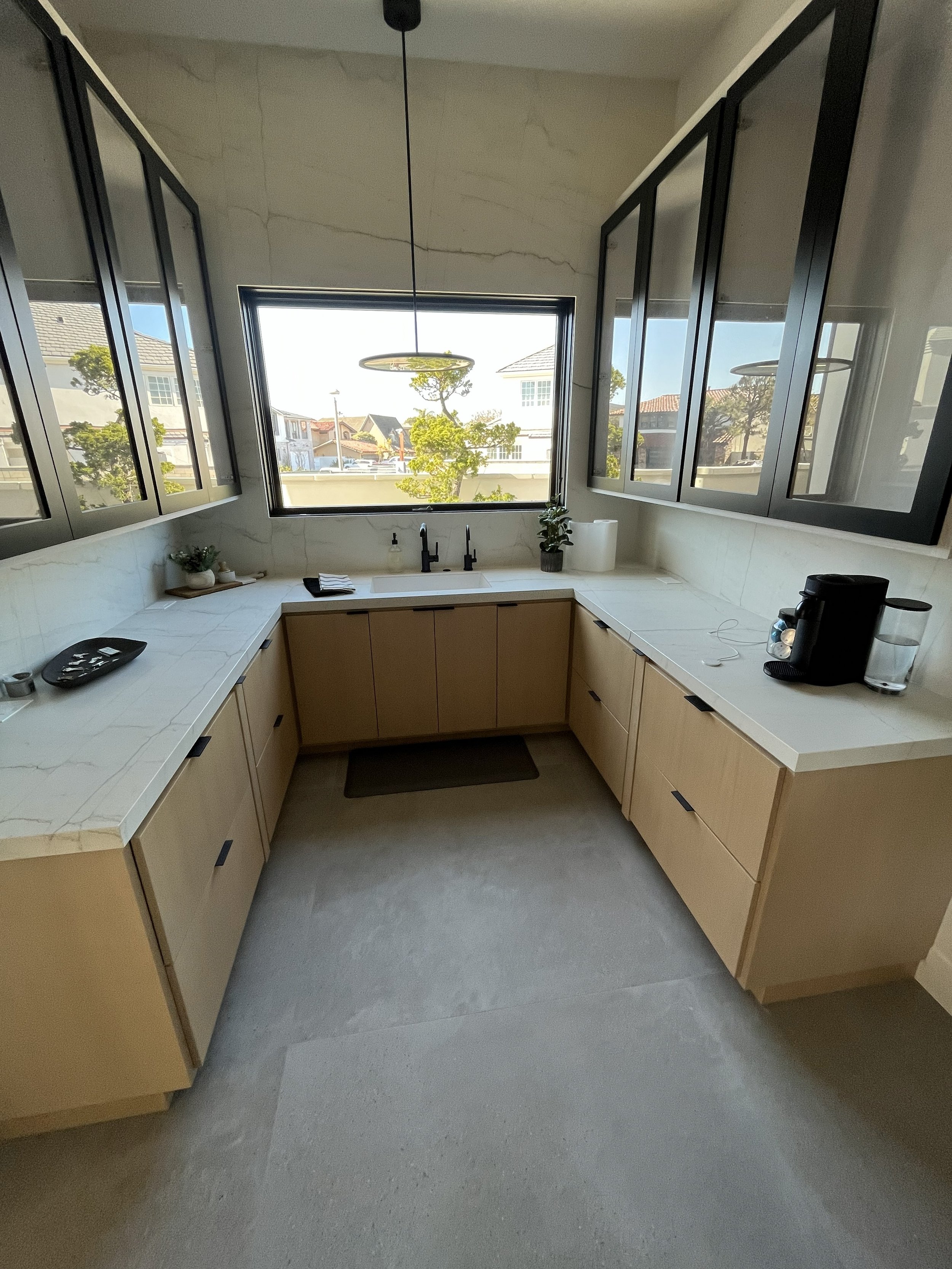 Modern kitchen with beige cabinets, a white marble countertop, and large black-framed windows overlooking a neighborhood with trees and houses.