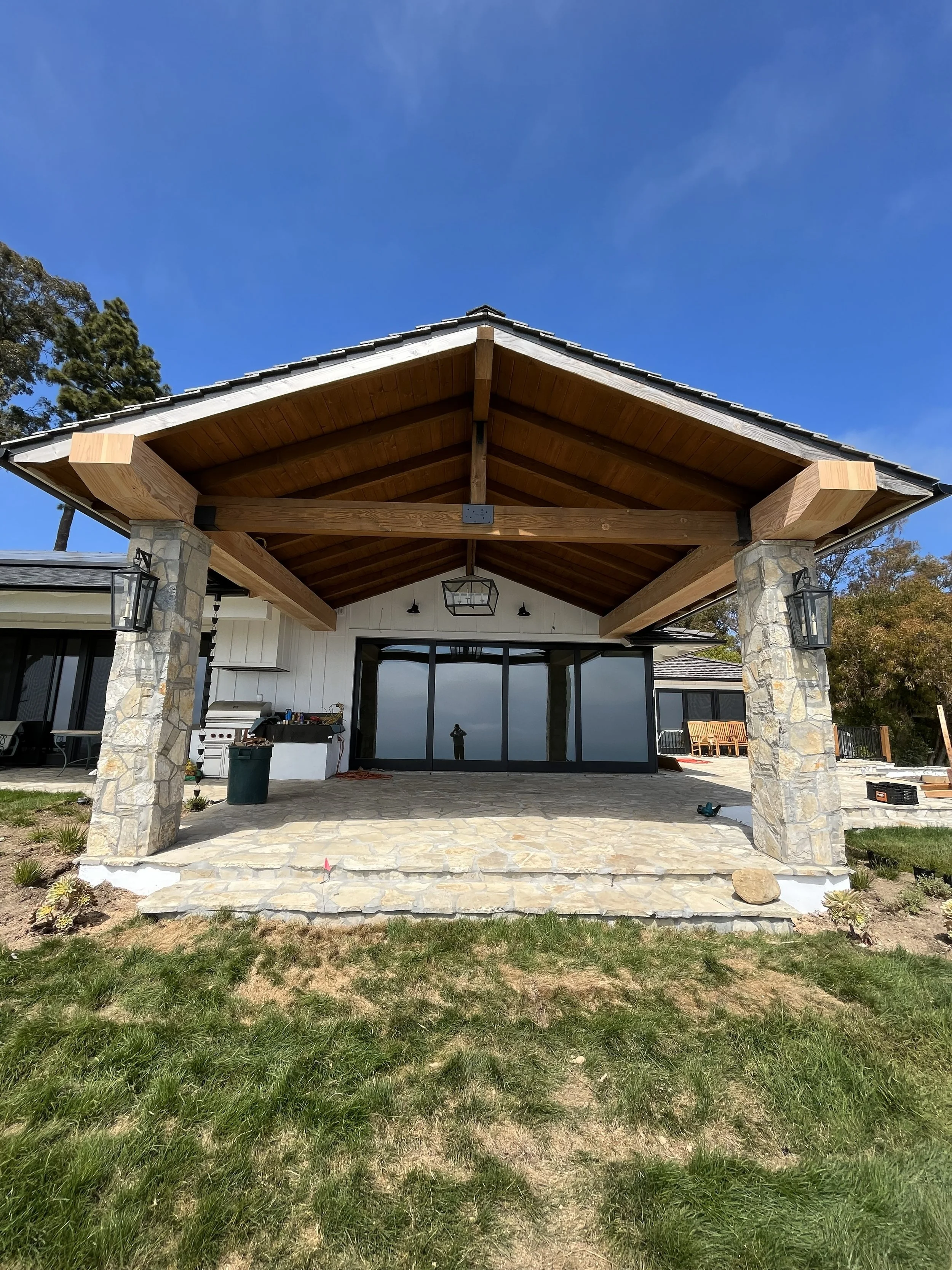 Construction site of a house patio with stone pillars and wooden roof structure, overlooking a grassy yard and a glass door.
