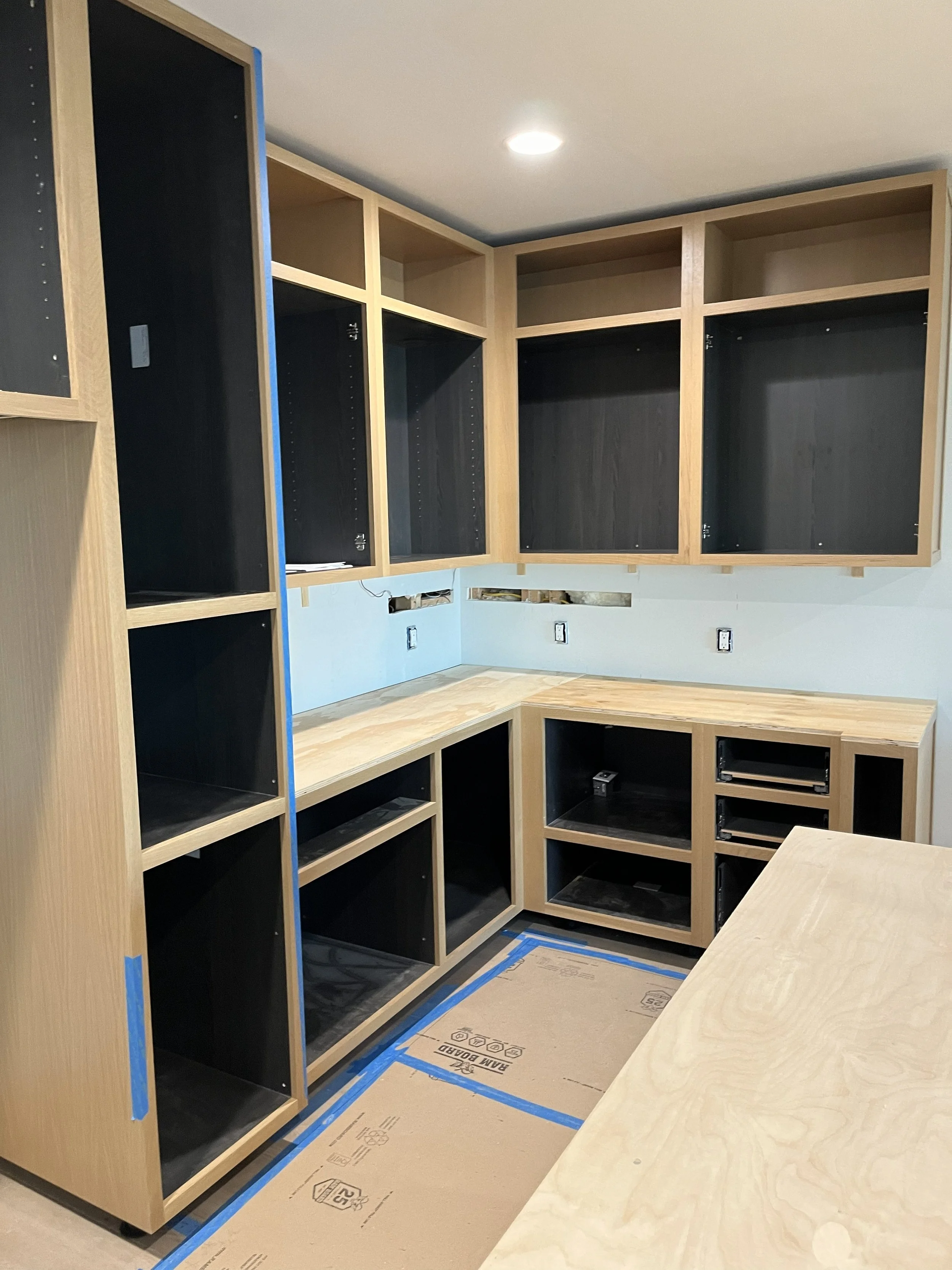 A kitchen or utility room under construction with wooden cabinets, some with black backing, and a wooden countertop. The floor is protected with cardboard and painters tape, and there are multiple wall outlets.