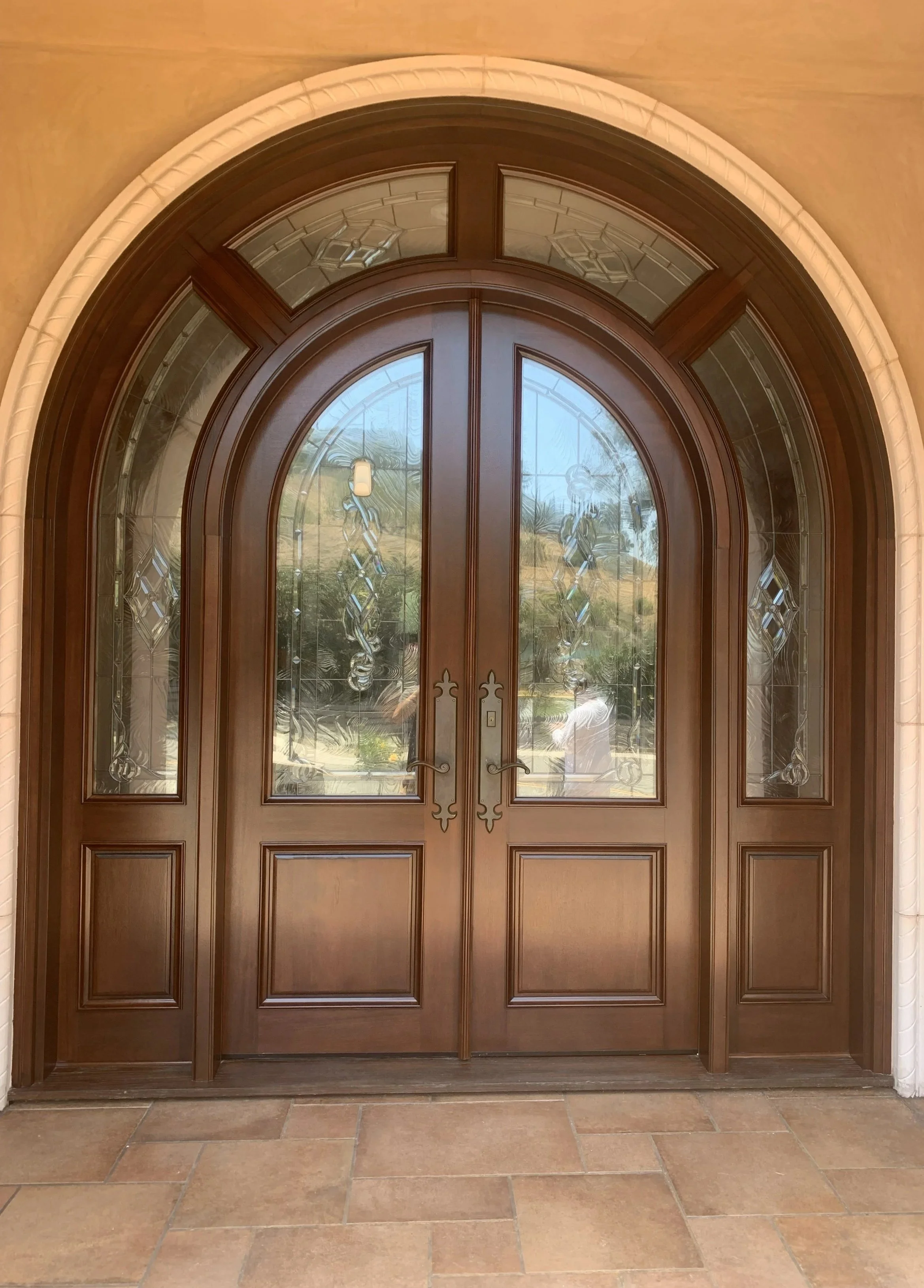 Double brown wooden doors with decorative glass panels and handles, set in an arched doorway with a tiled floor outside.