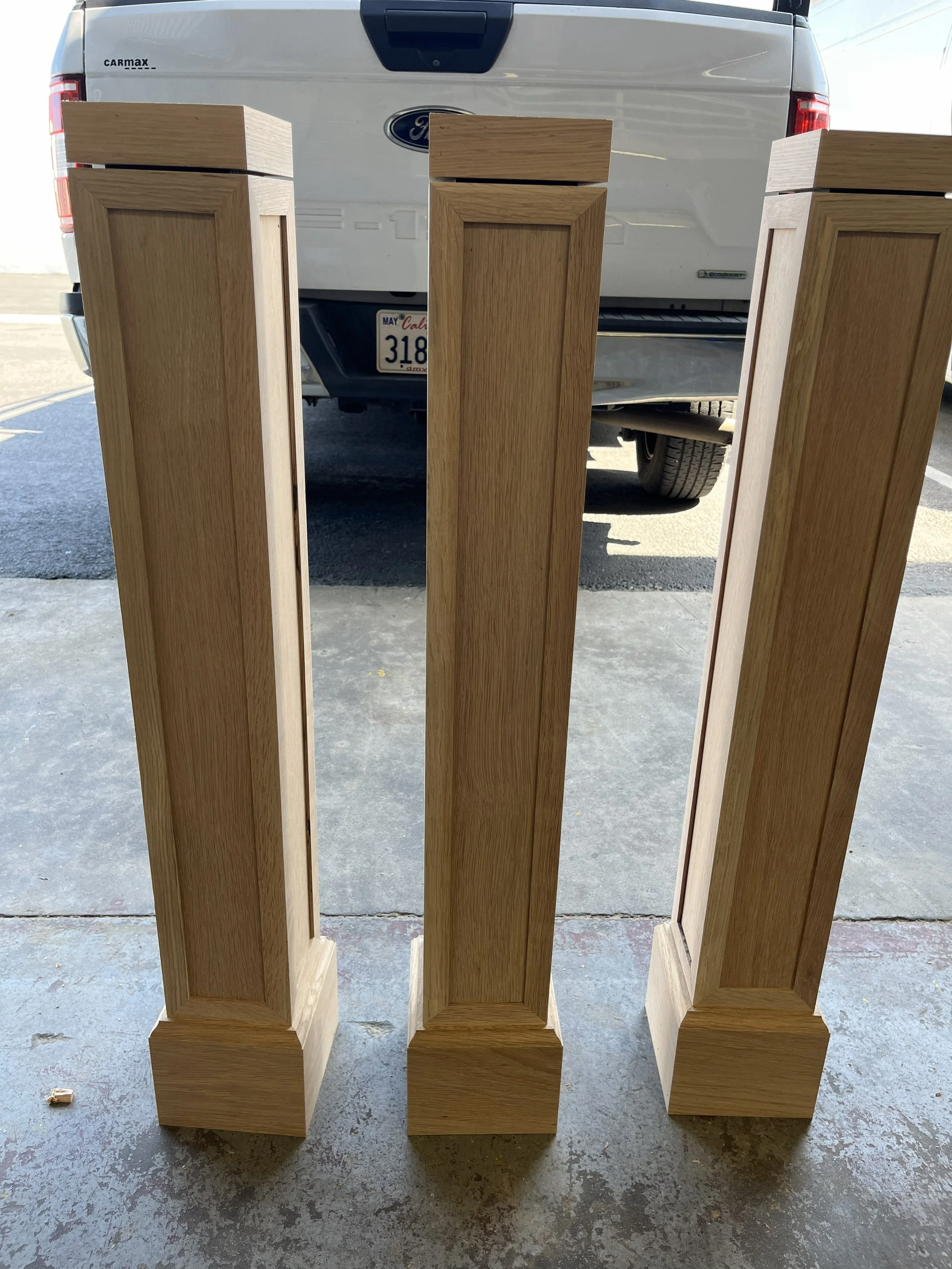 Three tall wooden cabinet sides with detailed paneling, standing upright on concrete floor outside a garage, with a white Ford pickup truck in the background.