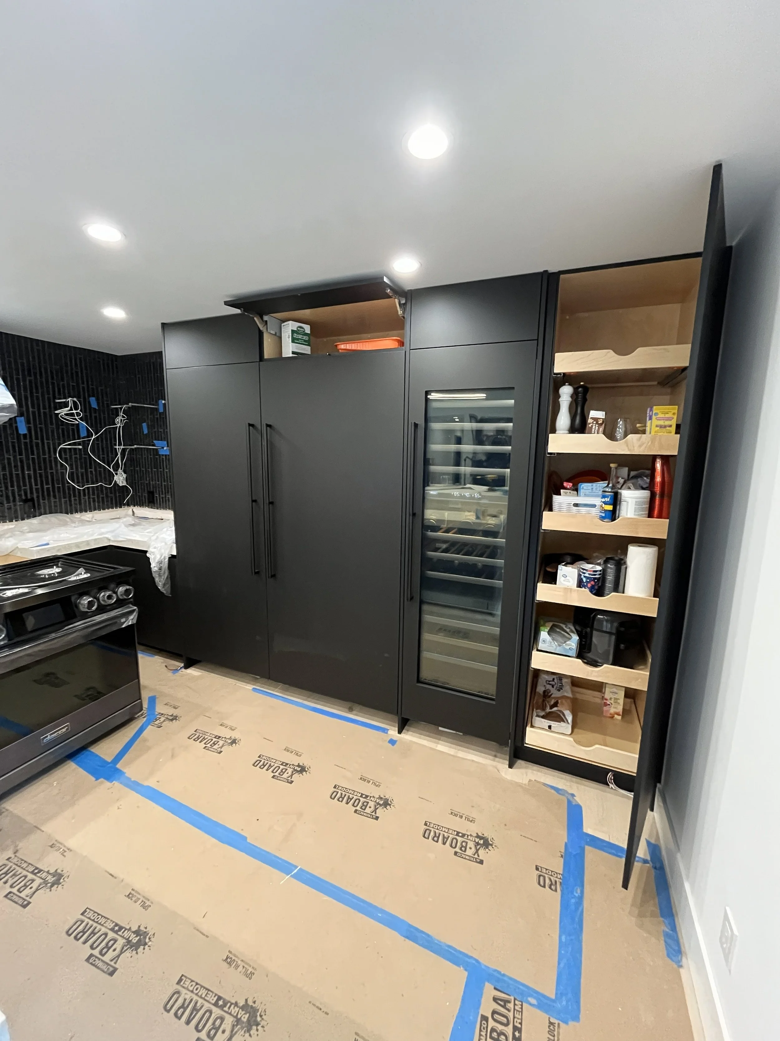 Kitchen with black cabinetry, a wine refrigerator, open shelves with various items, and a black stove. The floor is covered with brown protective paper and blue painter's tape.