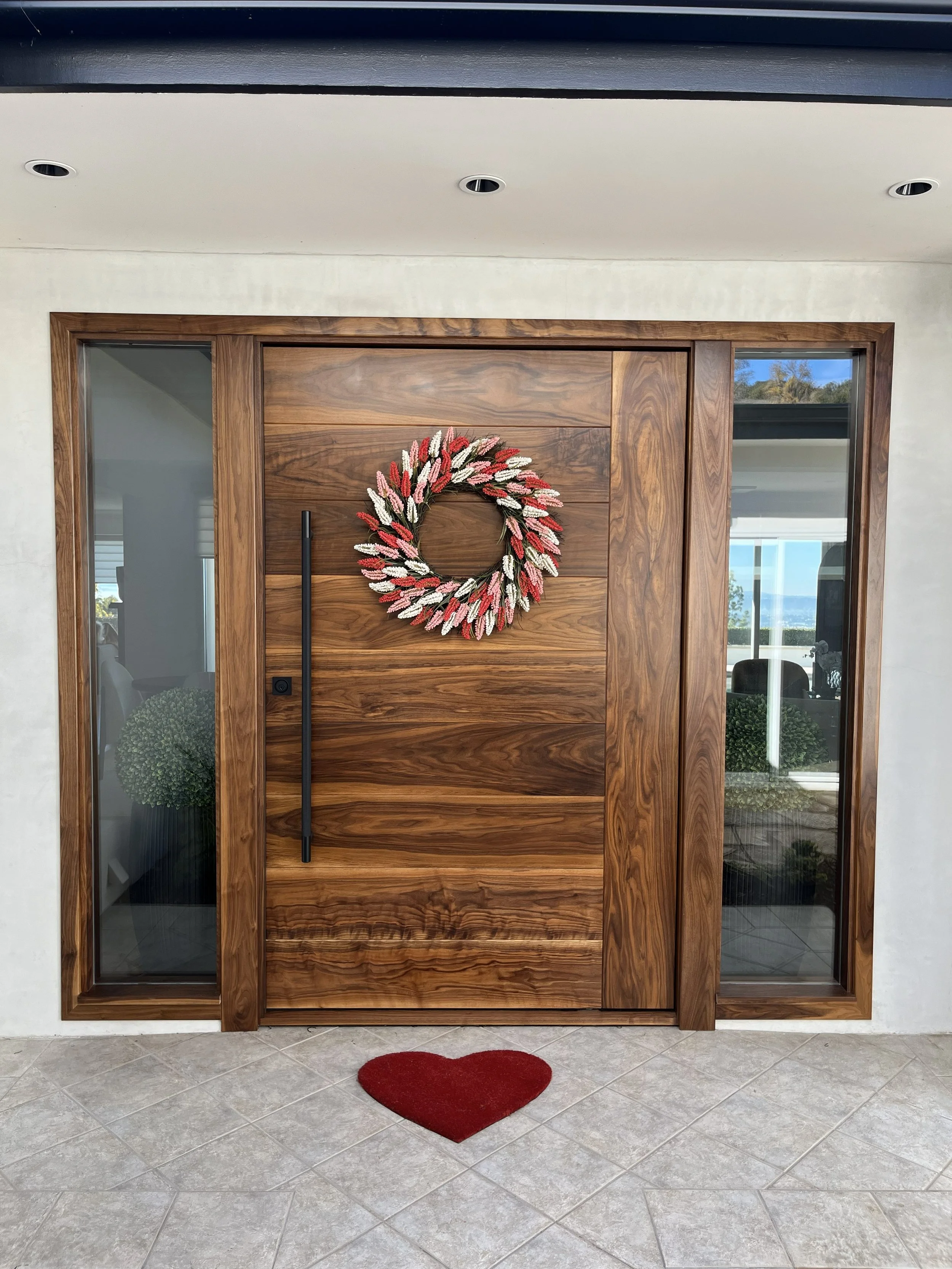 Front door with a wooden finish, decorated with a heart-shaped doormat and a wreath made of pink, red, and white flowers, flanked by side glass panels.
