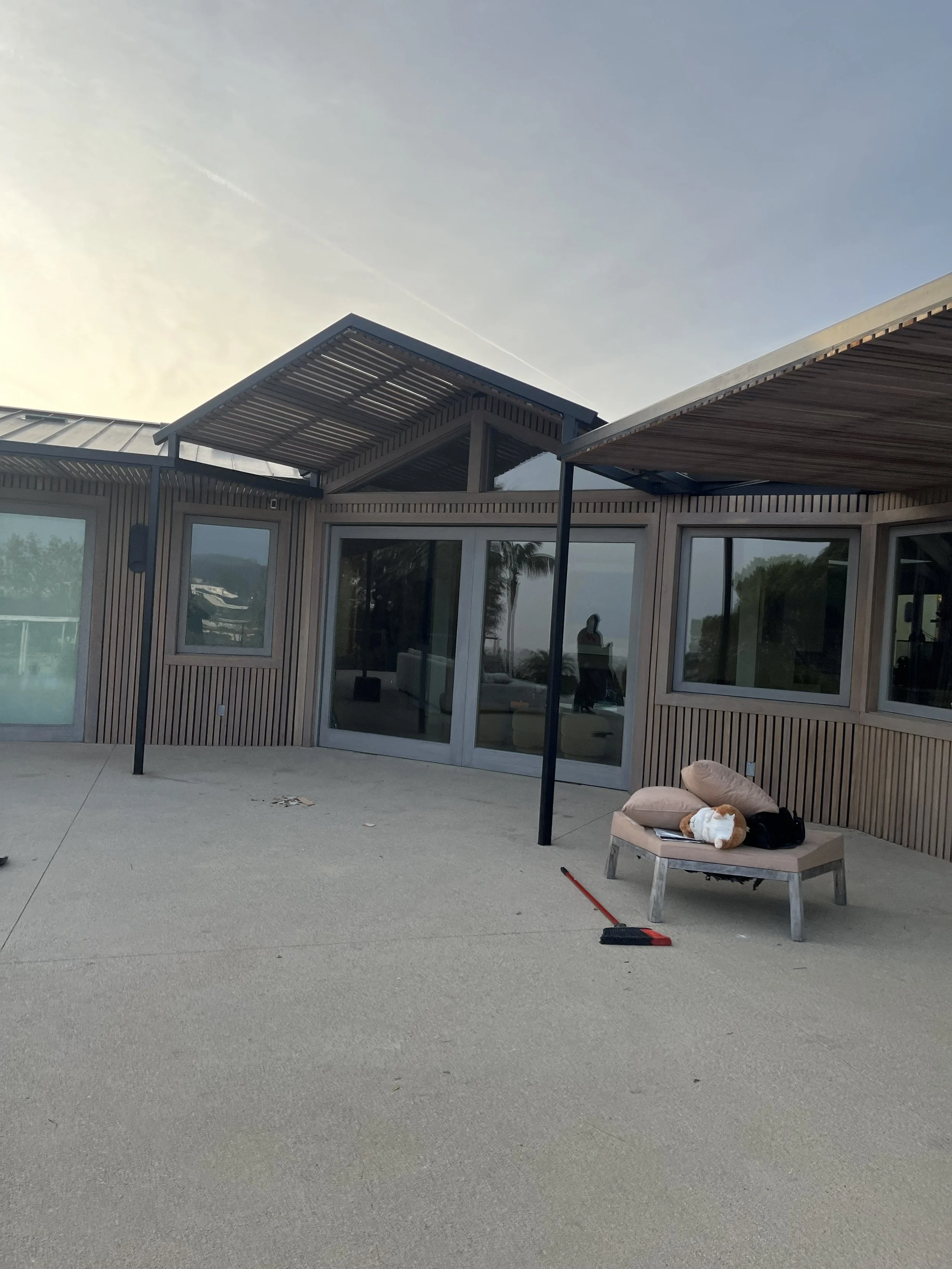 Modern house exterior with large glass sliding doors, deck furniture with cushions, and a broom on the ground during sunset.