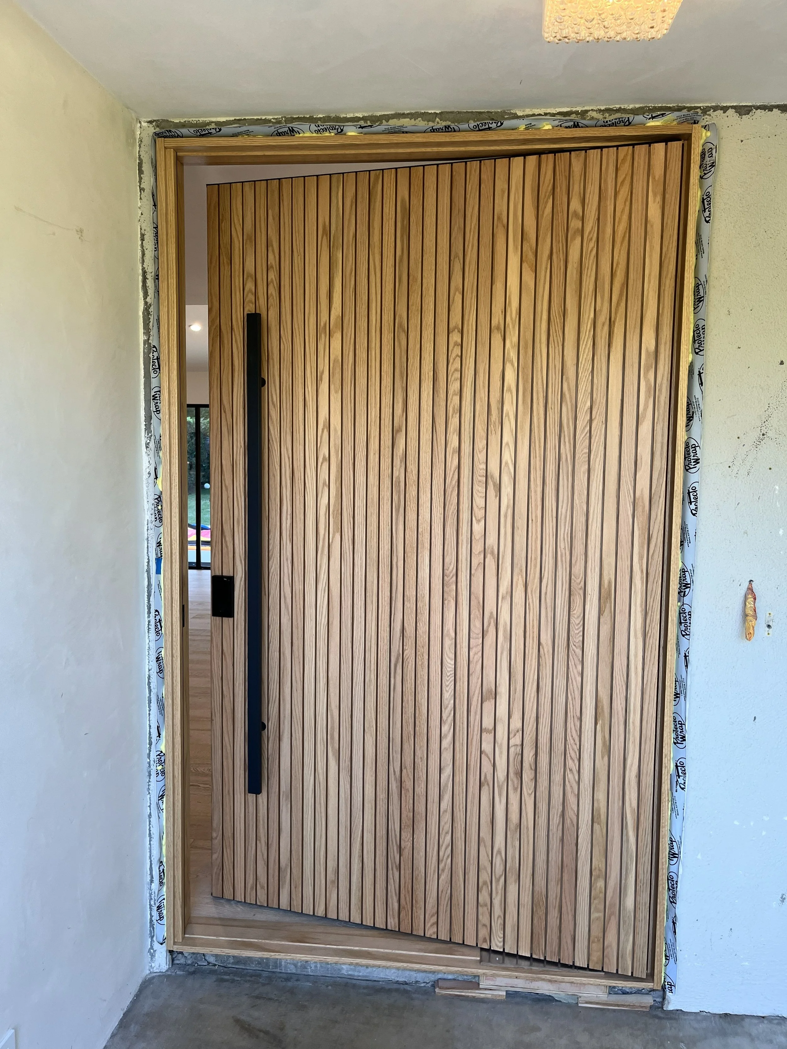 Wooden exterior door with a vertical slat design, black handle, and unfinished framing, part of a building under construction or renovation.