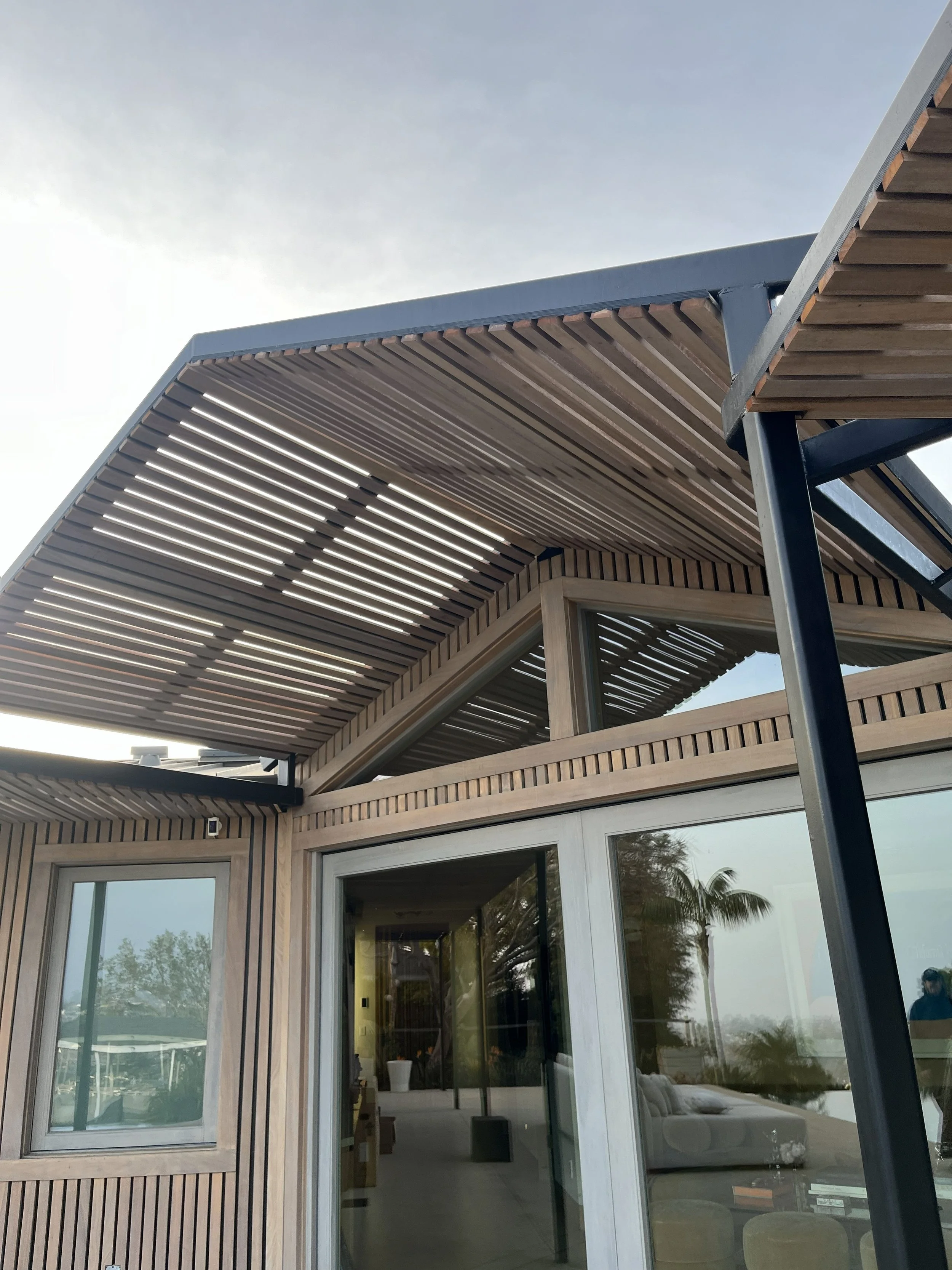 Close-up view of a modern house exterior with wooden slats and large glass sliding doors, featuring an overhanging roof with a slatted design, and reflected trees in the glass.