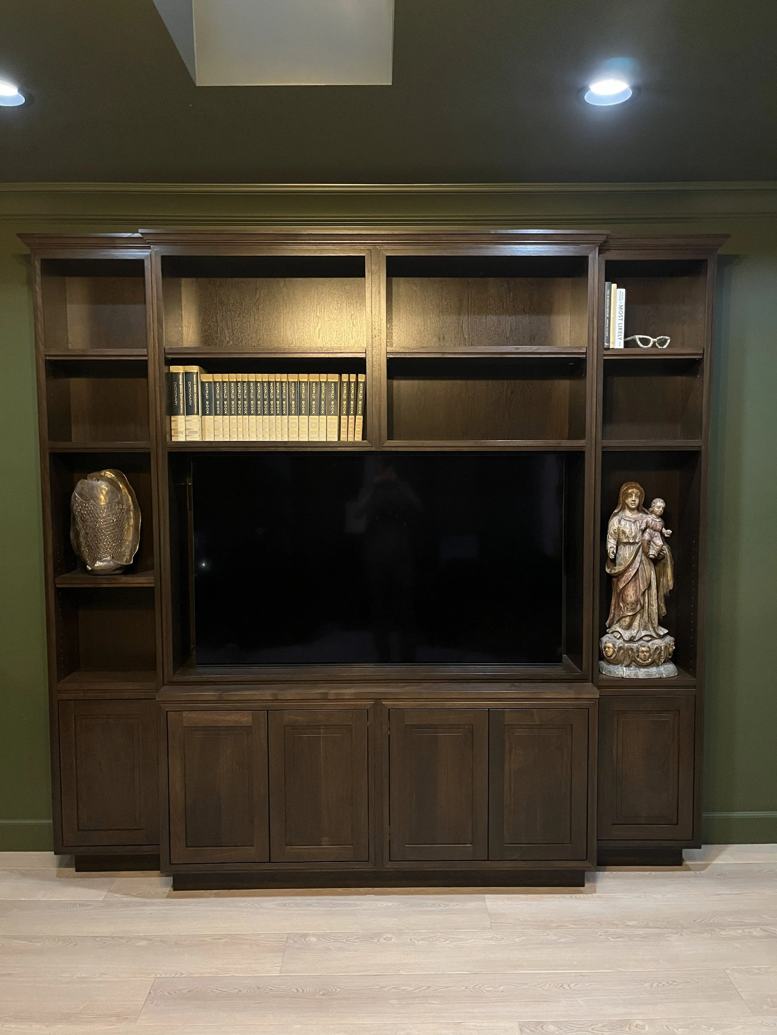 A dark wood entertainment center with a large flat screen TV in the middle, shelves above and below, and decorative items on the sides, including a decorative owl and a religious statue of a woman holding a child.