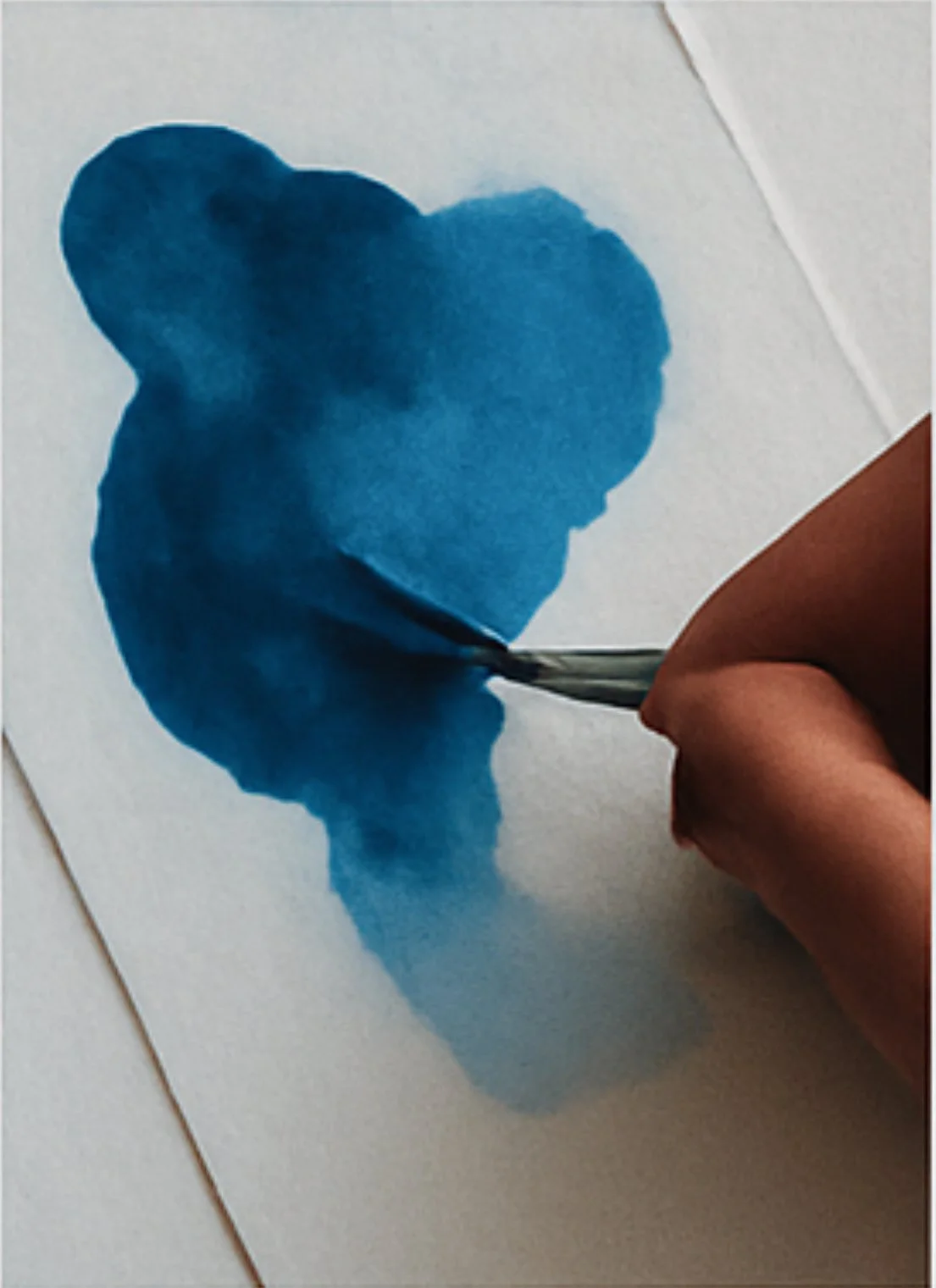 A hand using a paintbrush to create an abstract watercolor painting in blue on white paper.