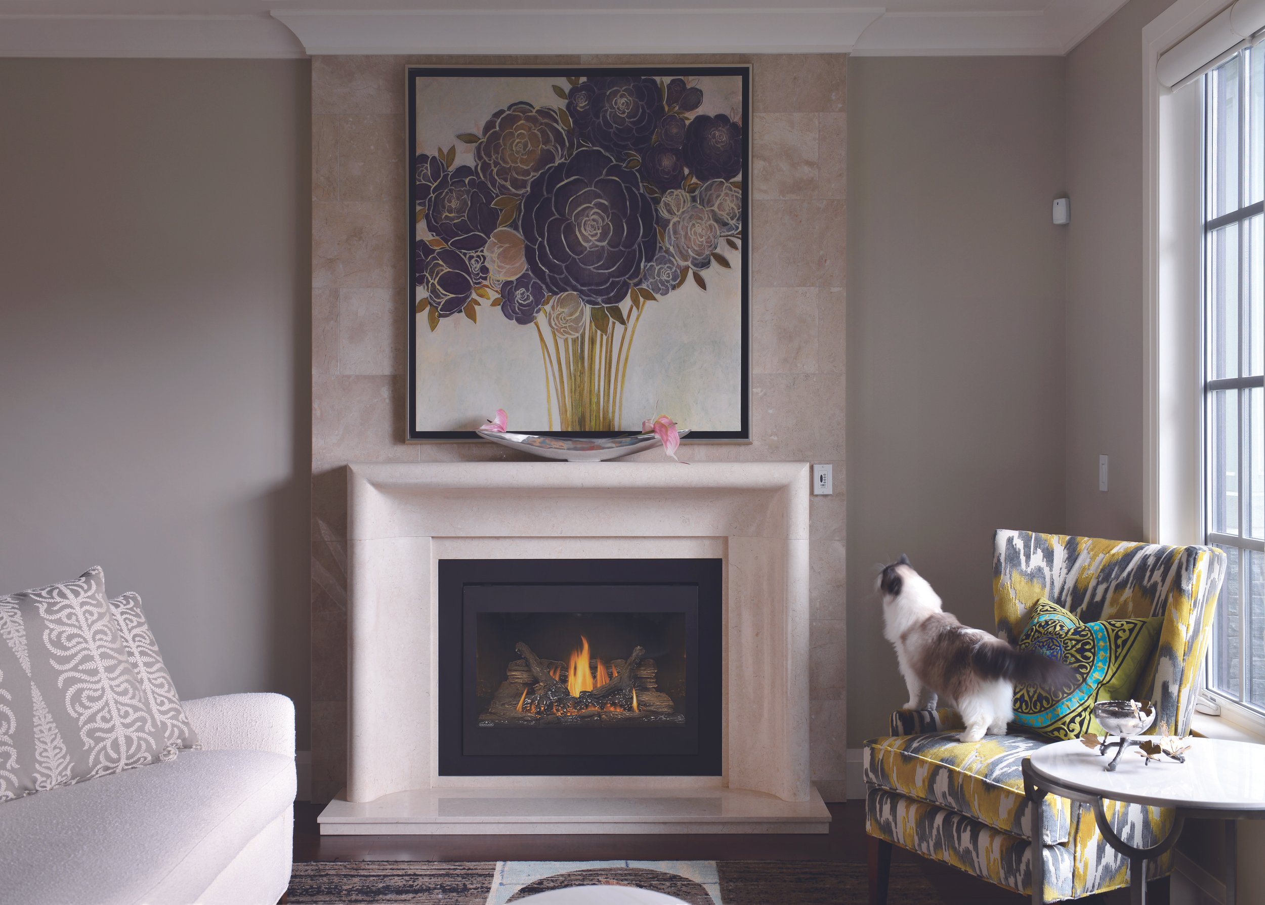Living room with a fireplace, a framed floral art piece above it, a colorful armchair with a black and white cat sitting on it, a side table with decorative items, and a window letting in natural light.