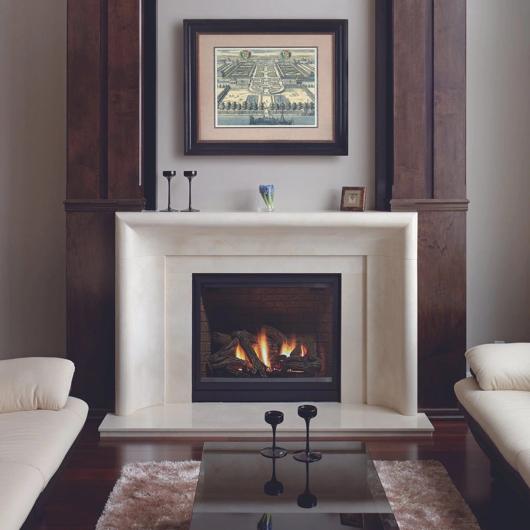 Living room with a white fireplace, framed artwork above, fireplace with burning logs, dark wood paneling, two white sofas, glass coffee table with two black candle holders, area rug.