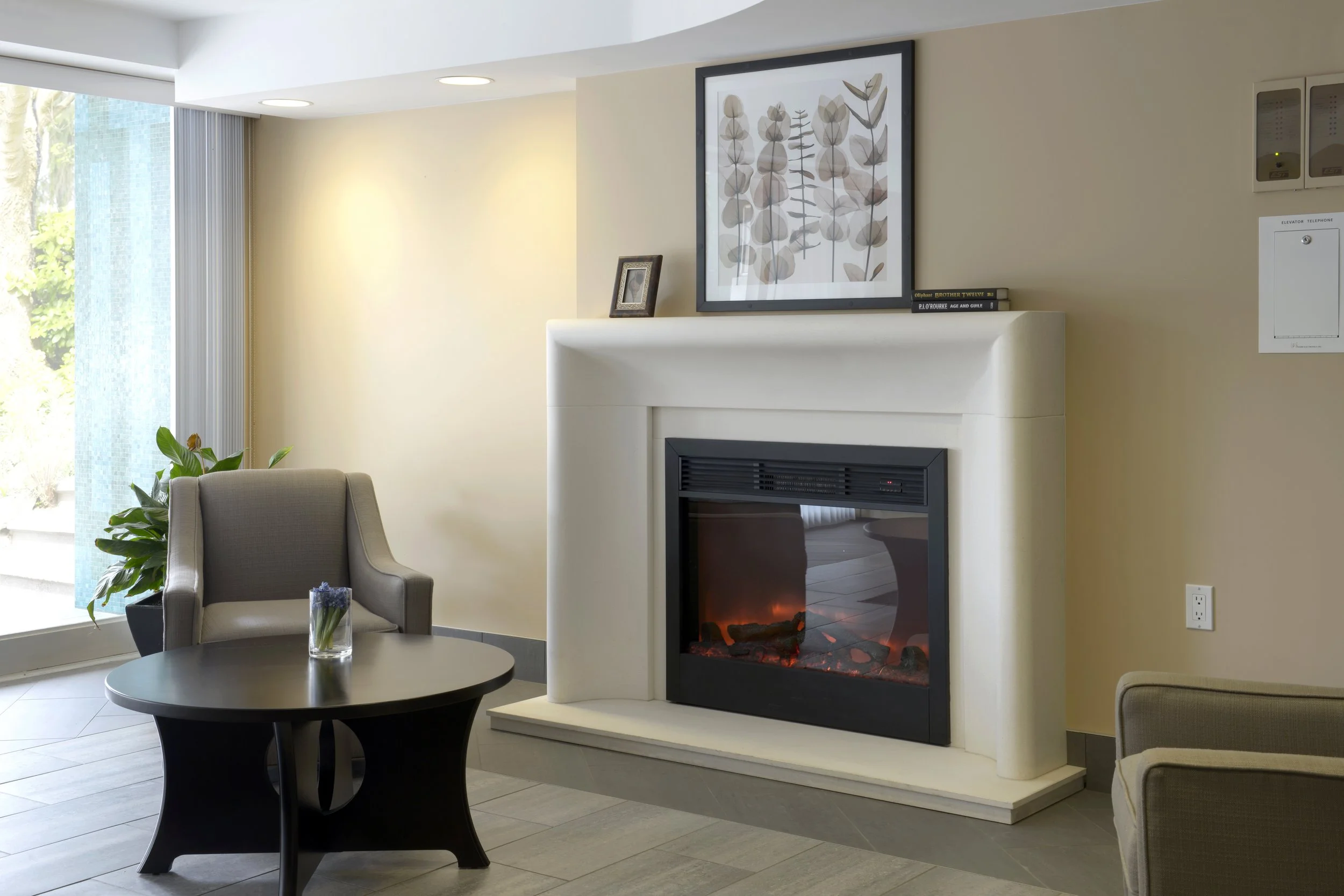 Living room with fireplace, gray armchair, black coffee table, potted plant, large window with vertical blinds, and framed artwork above the fireplace.