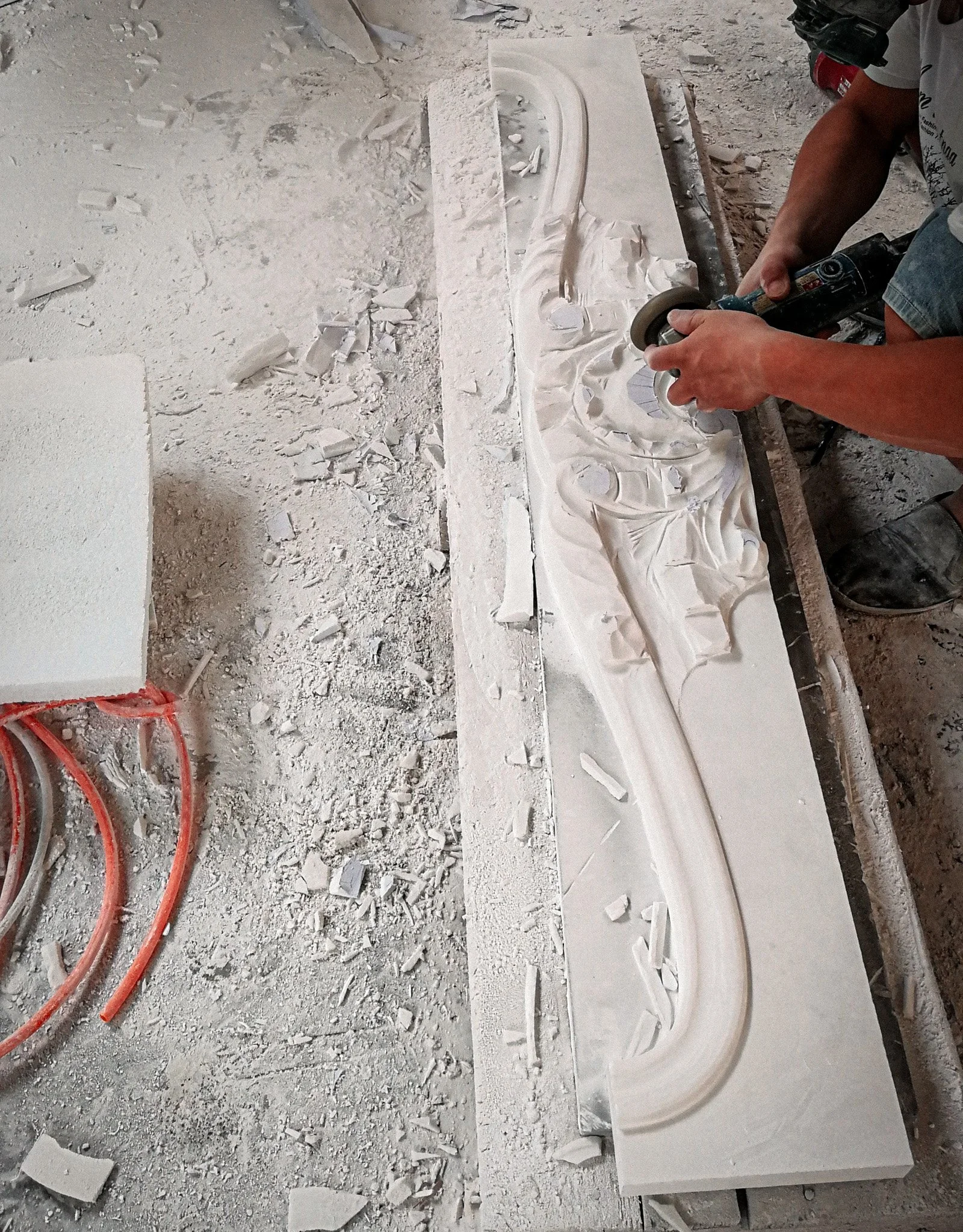 A person is carving a decorative design into a large white stone slab with a power tool, surrounded by stone dust and debris.