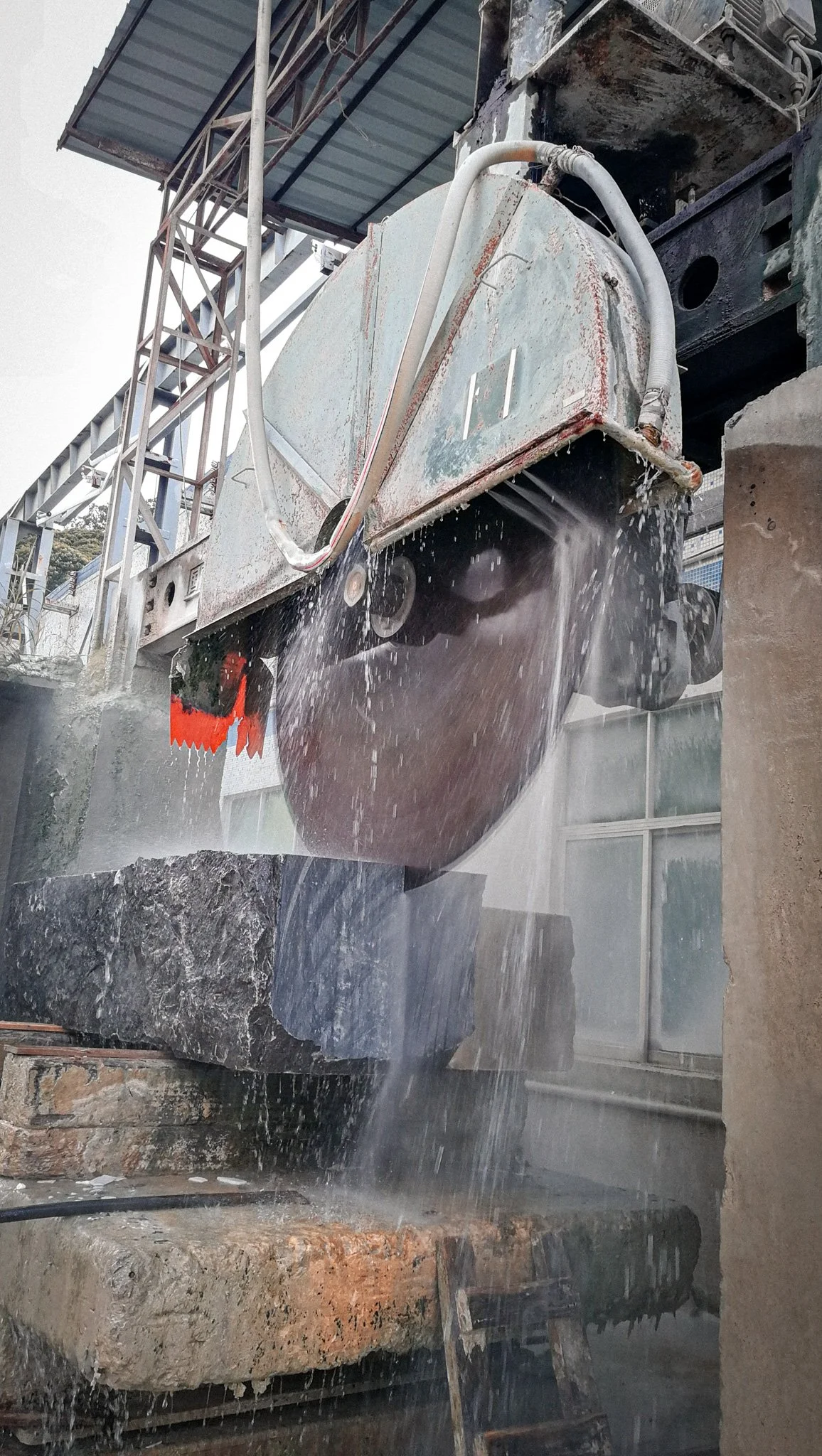 A large circular saw cutting through a stone block outdoors, with water splashing to cool the blade, under a metal roofed structure.