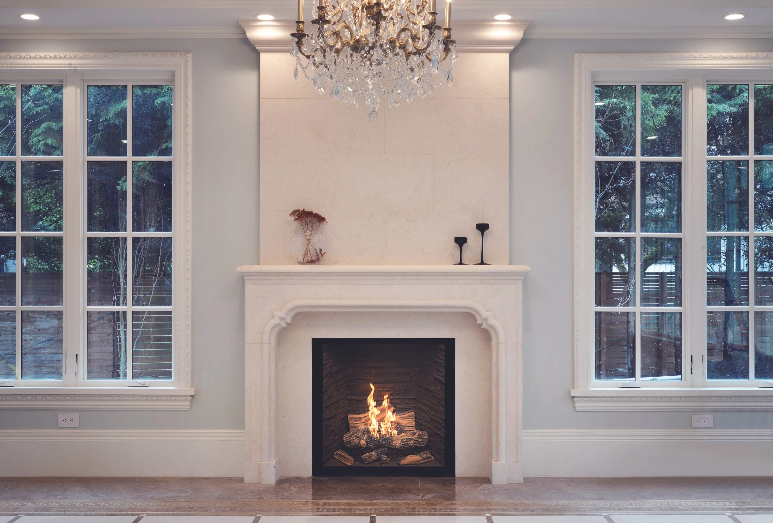 Elegant living room with a white fireplace, two large windows, a chandelier, and decorative items on the mantel.
