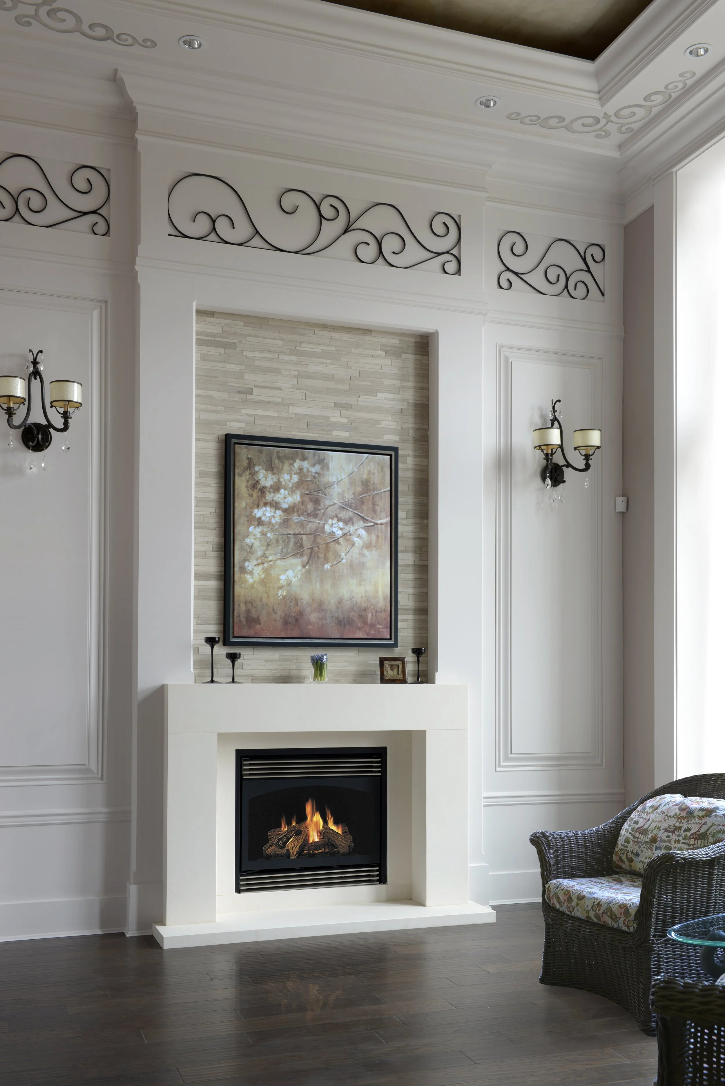 A living room with a white fireplace with a fire burning inside, framed artwork above it, and candles and a small framed picture on the mantle. The room has white walls with decorative molding and black metal wall sconces with candle-shaped lights. There is a brown wicker armchair with floral cushion on the right and a glass table in front.