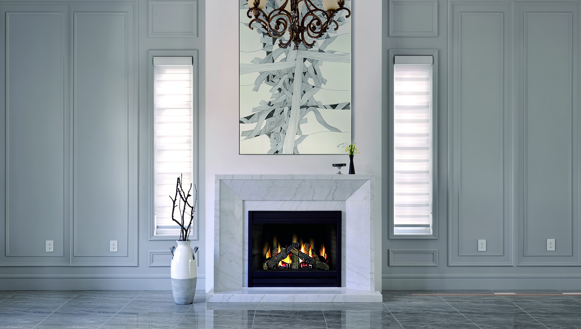 Living room with a white marble fireplace, two tall windows with blinds, a chandelier, abstract artwork above the fireplace, and a white vase with decorative branches to the left of the fireplace.