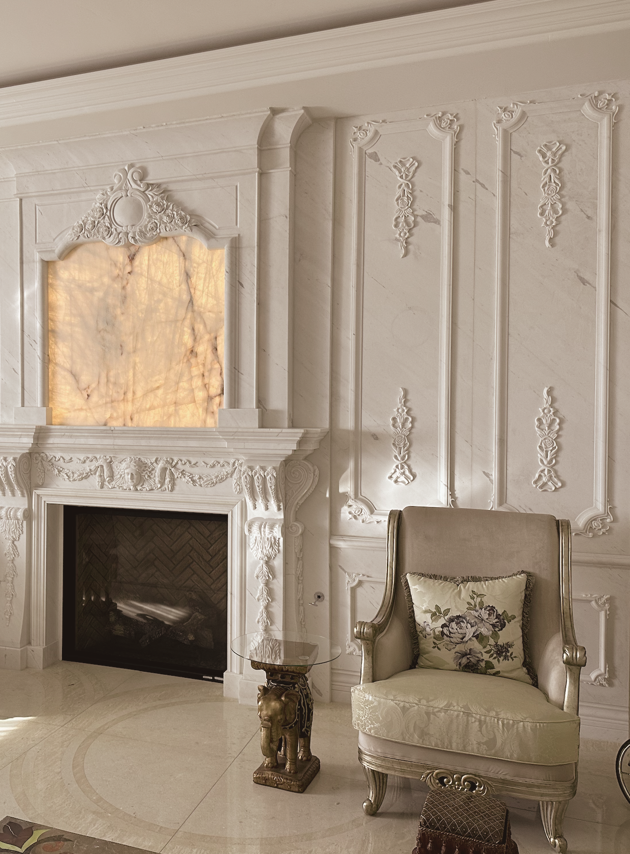 Elegant living room corner with a white ornate fireplace, beige armchair with a floral pillow, a small glass-topped table on an elephant-shaped base, and decorative wall panels.