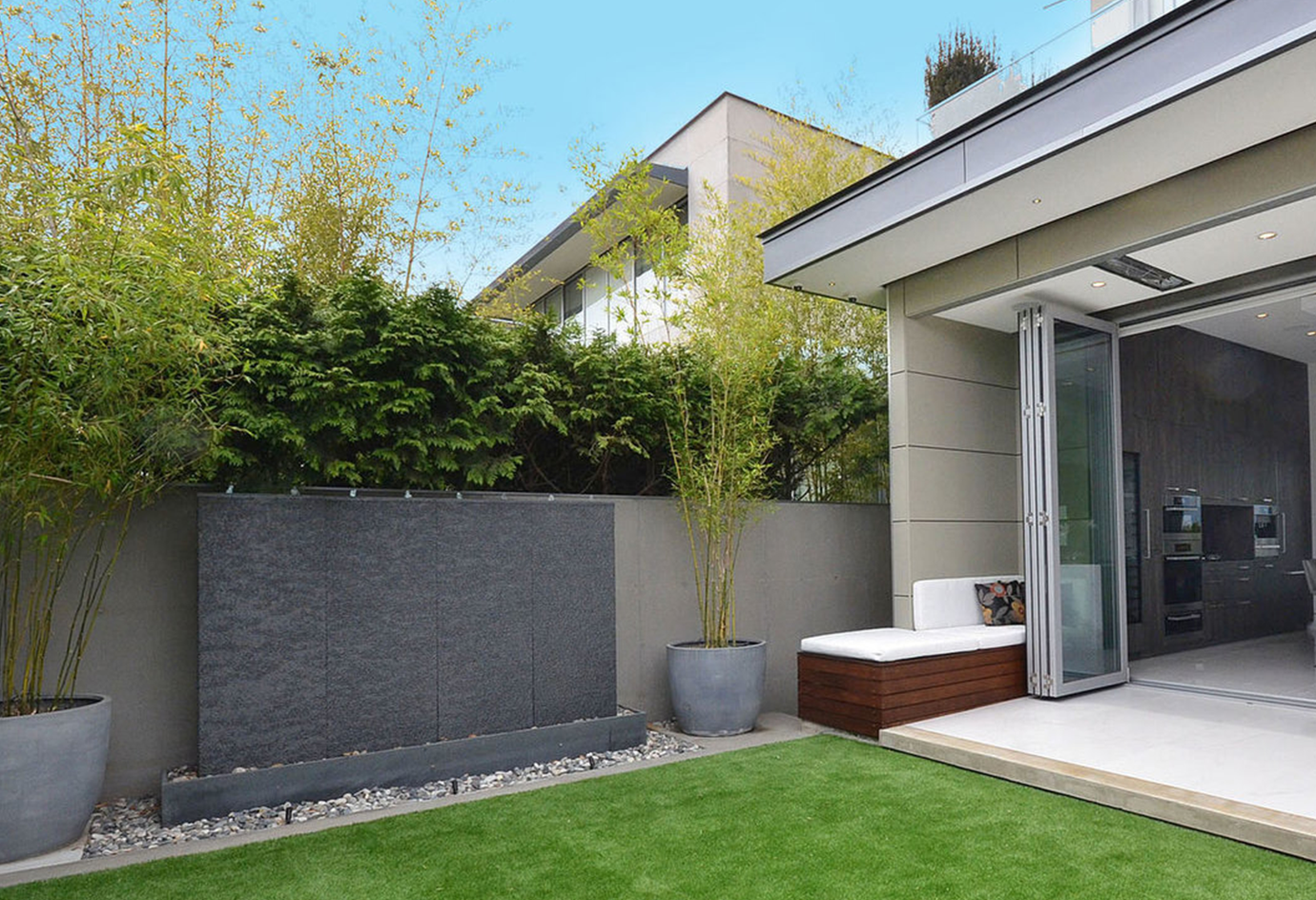 Modern backyard patio with green grass, potted plants, and a bench with a cushion next to a sliding glass door leading inside a house.