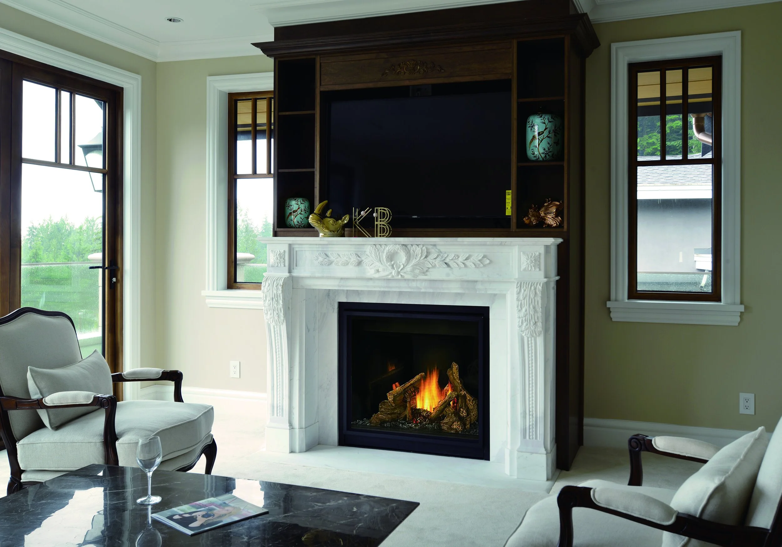 Living room with a white marble fireplace, turned on with a fire, surrounded by armchairs, a coffee table with a magazine and a wine glass, decorated shelves, and large windows showing greenery outside.