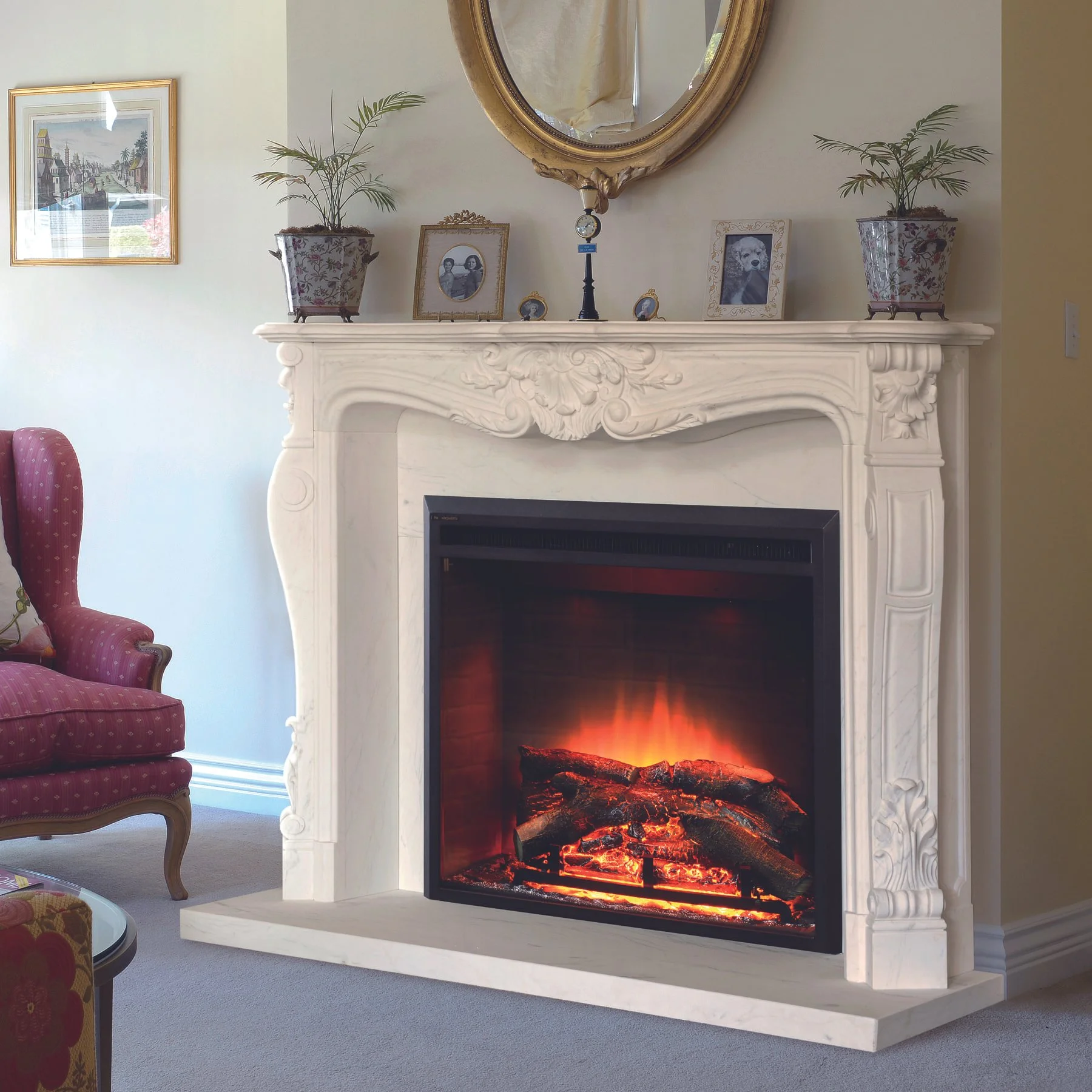 Living room fireplace with a lit electric fire, ornate white mantel decorated with framed photos and potted plants, red upholstered armchair to the left, and framed artwork on light-colored walls.