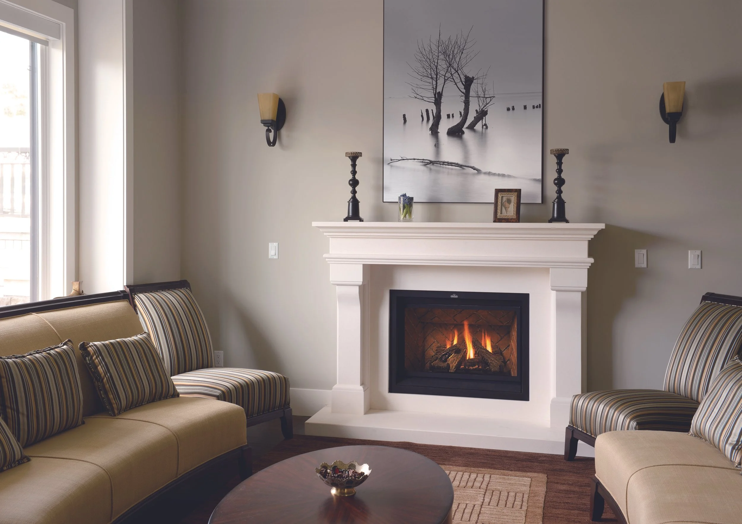 Living room with beige sofa chairs, striped cushions, a white fireplace with a fire burning, a large black-and-white artwork of trees and water above the fireplace, two candle holders, a small framed photo, a small plant, a round wooden coffee table with a decorative bowl, and wall sconces on a light-colored wall.