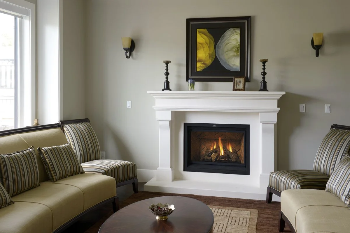 Living room with a fireplace, yellow and striped armchairs, a round coffee table, wall sconces, and a framed painting above the fireplace.
