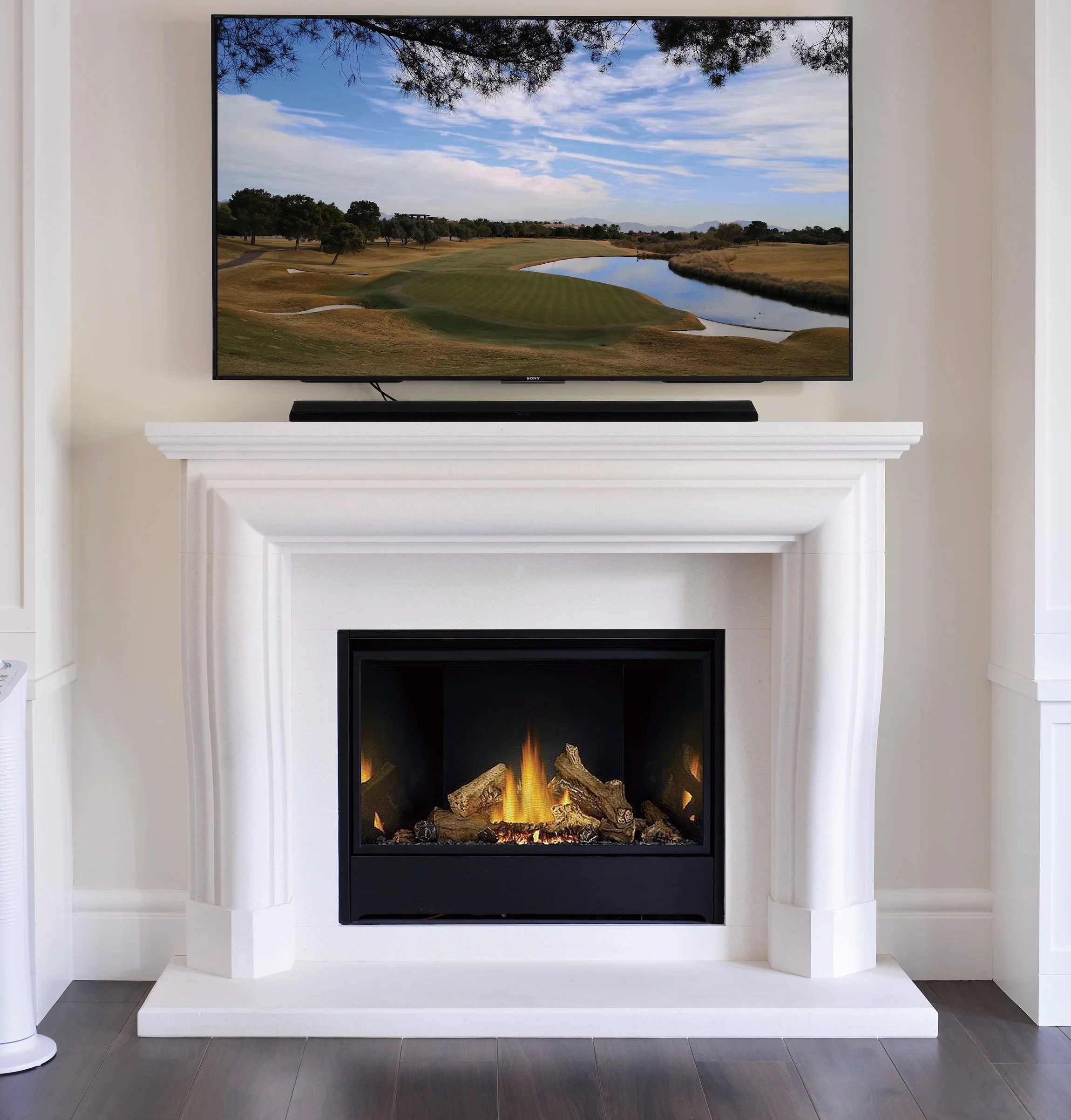 A living room with a white fireplace and a mounted flat-screen TV displaying a golf course landscape with trees, water, and a partly cloudy sky.