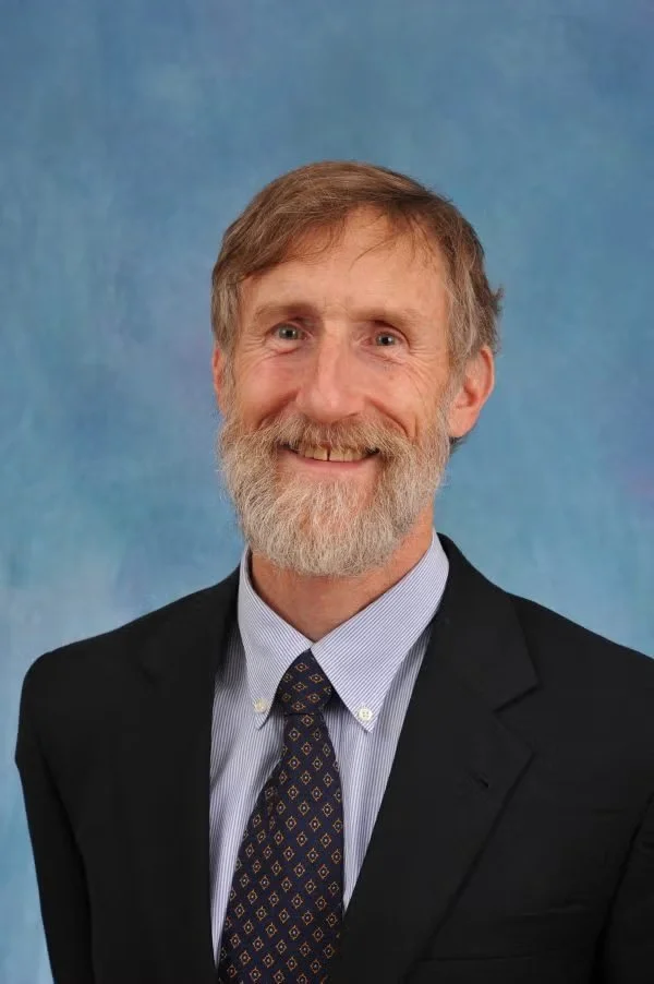 A middle-aged man with gray hair and a beard, wearing a dark suit, a light blue shirt, and a patterned tie, smiling in front of a blue gradient background.