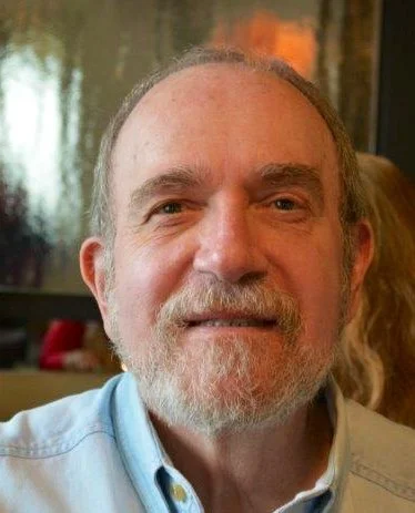 A middle-aged man with a beard and mustache smiling, wearing a light blue shirt, in an indoor setting with blurred background.