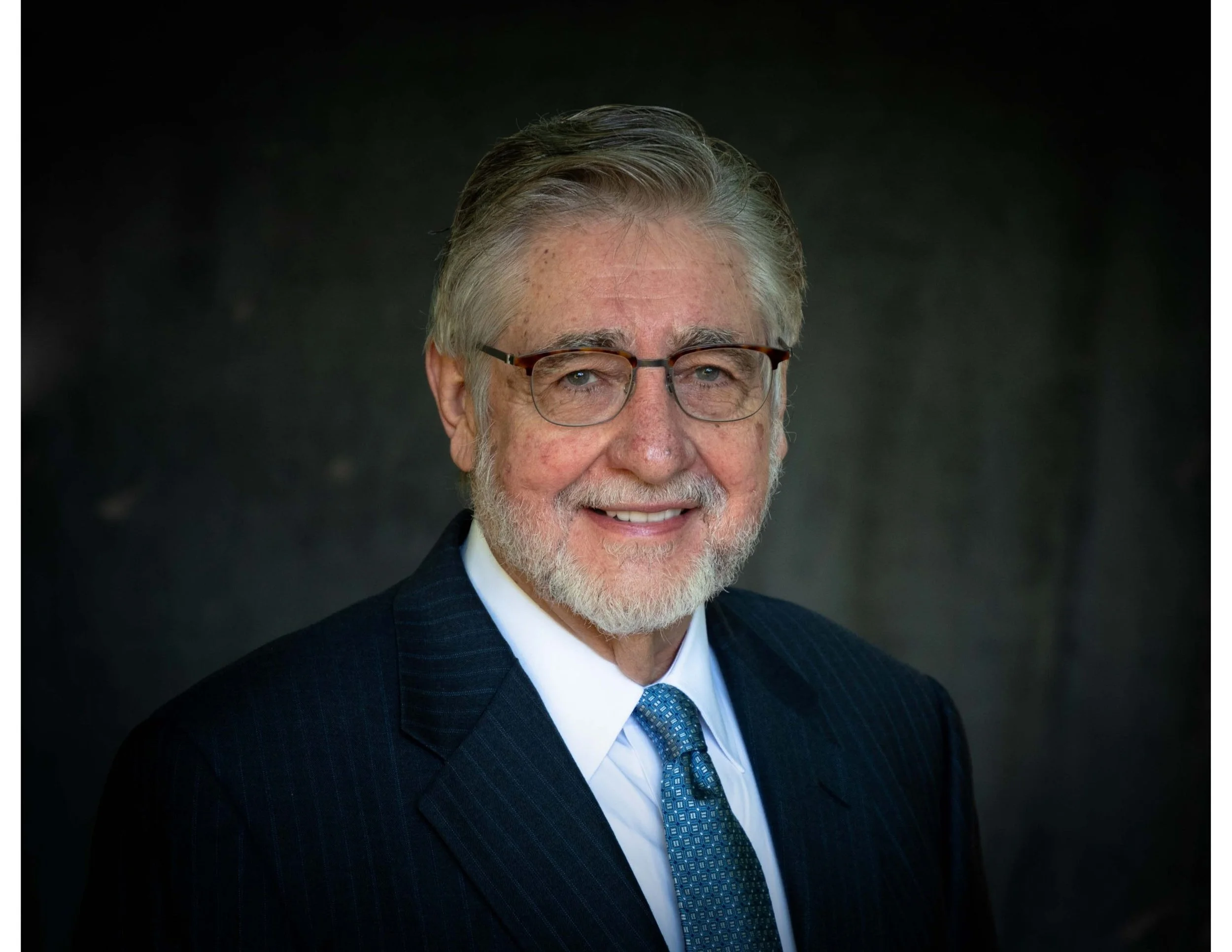 Senior man with glasses, gray hair, and a beard, wearing a dark pinstripe suit, white shirt, and blue patterned tie, smiling against a dark background.