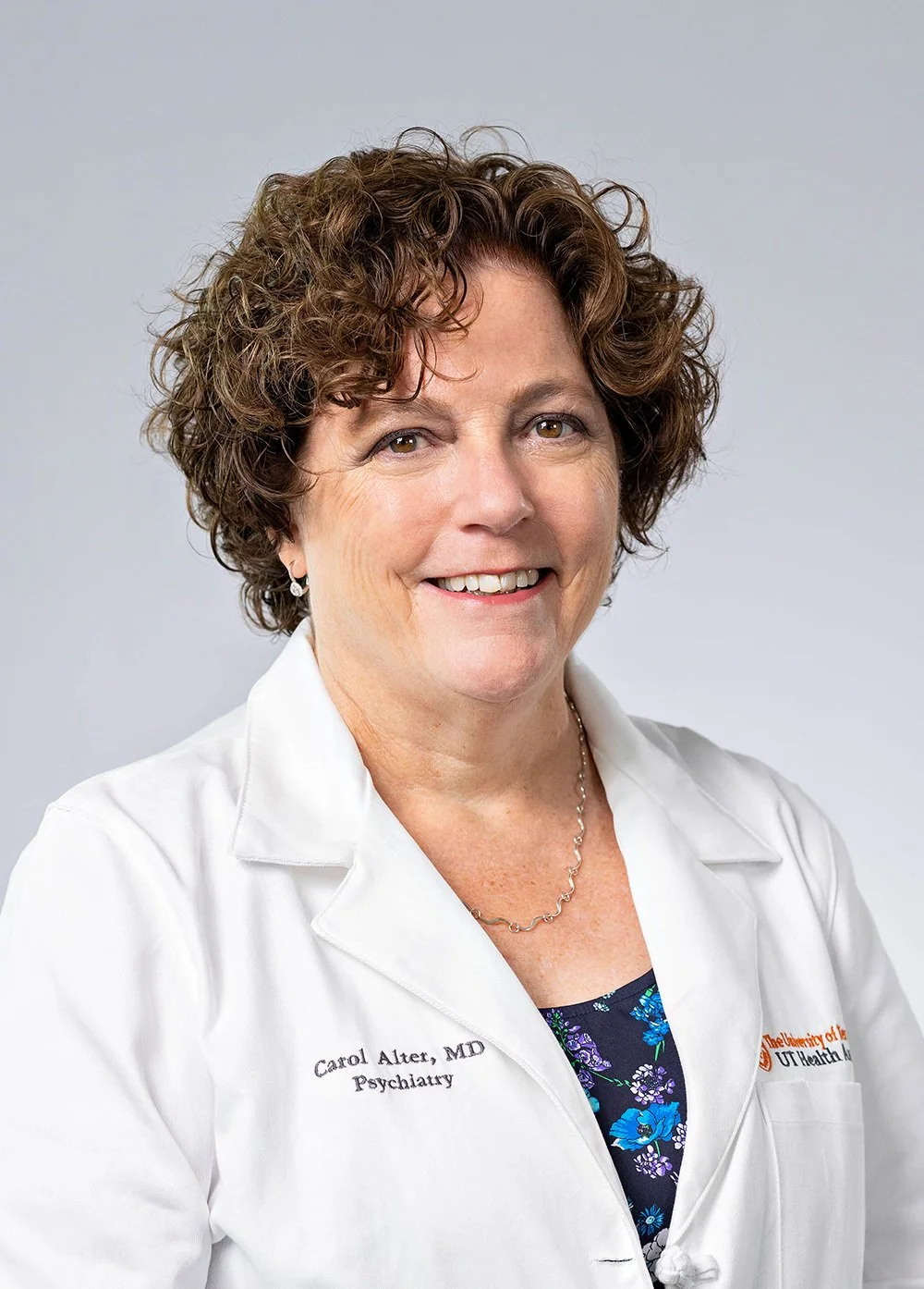 A smiling woman with short, curly brown hair wearing a white lab coat with her name and credentials embroidered on it. She is also wearing a blue floral blouse and jewelry, against a light gray background.