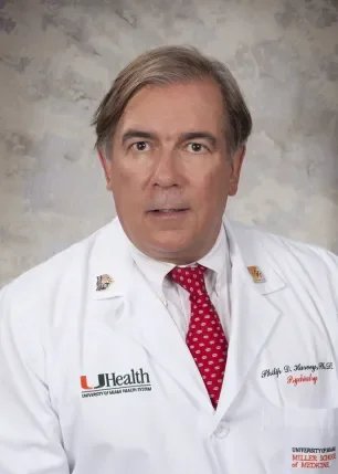 A middle-aged man wearing a white lab coat with the University of Miami Health logo, red polka dot tie, and a name tag, standing against a gray background.