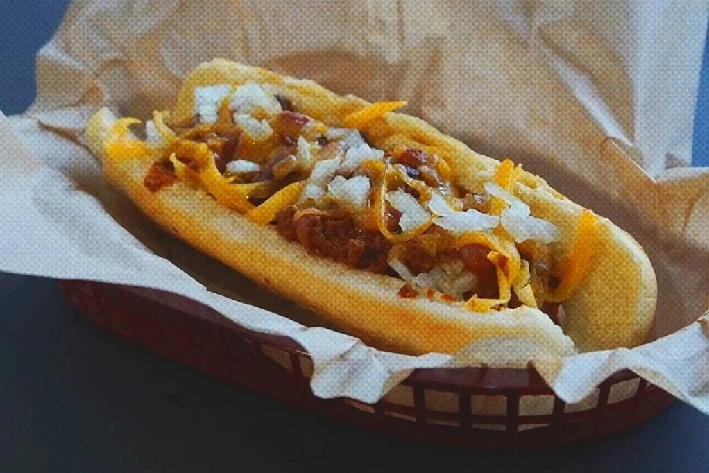 Hot dog topped with chili, shredded cheese, and chopped onions in a paper-lined basket.
